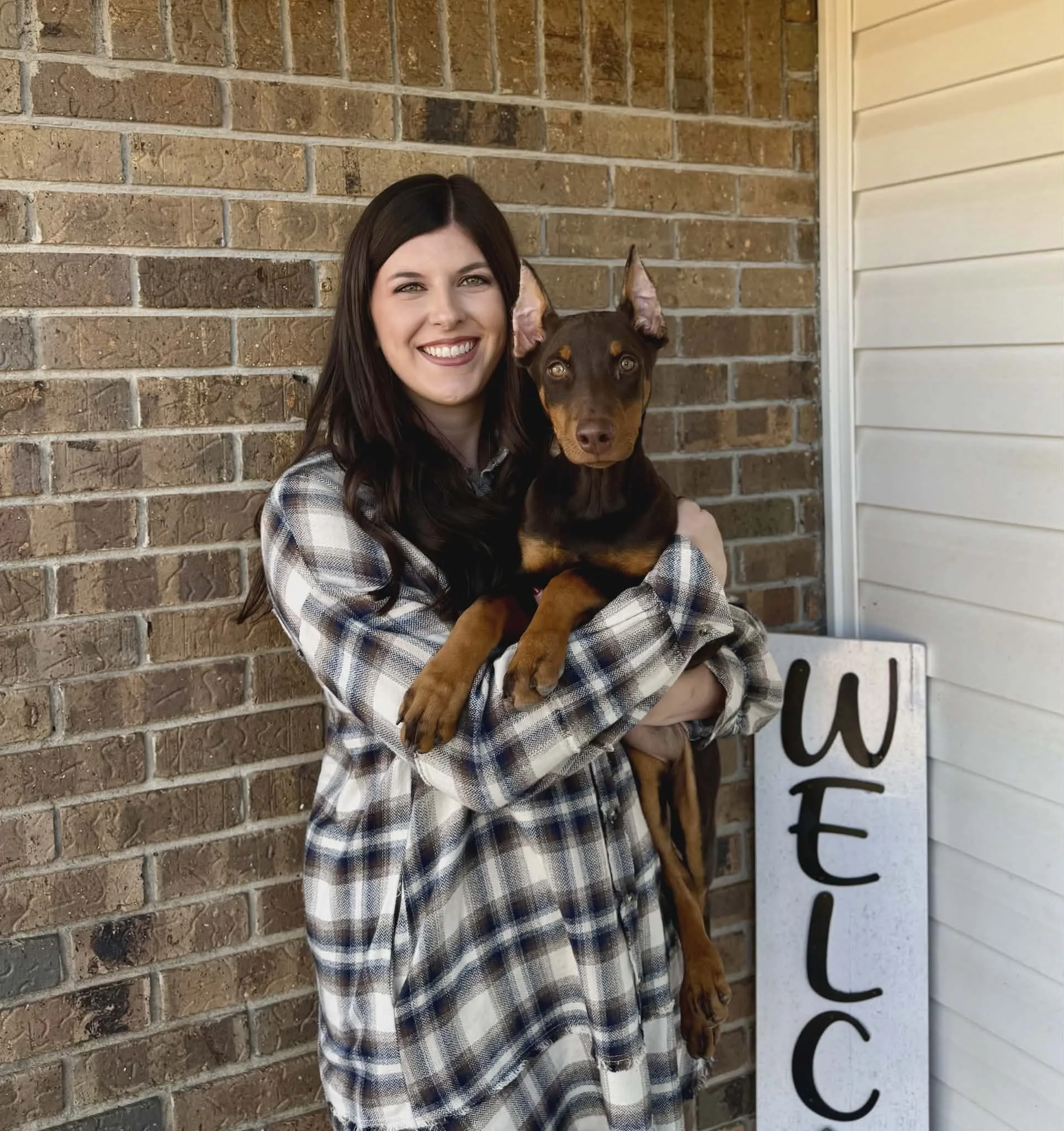 A young woman with long dark hair smiling and holding a Doberman puppy in front of a brick wall and cream-colored siding, with a welcome sign nearby.