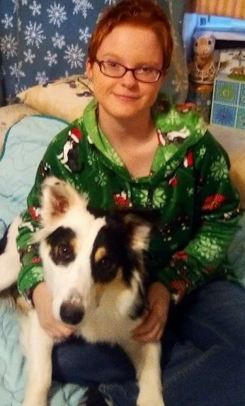 A person with short red hair and glasses sitting on a bed, wearing a green holiday sweater, holding a black, white, and brown dog.