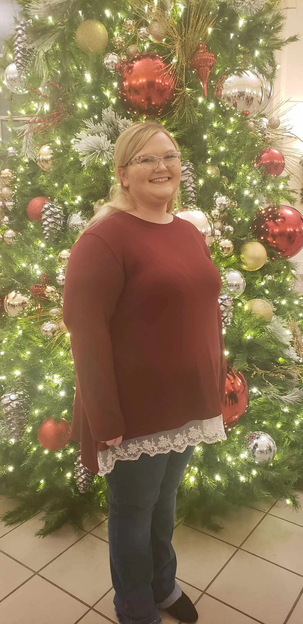 A woman standing in front of a decorated Christmas tree with lights and ornaments, smiling at the camera.