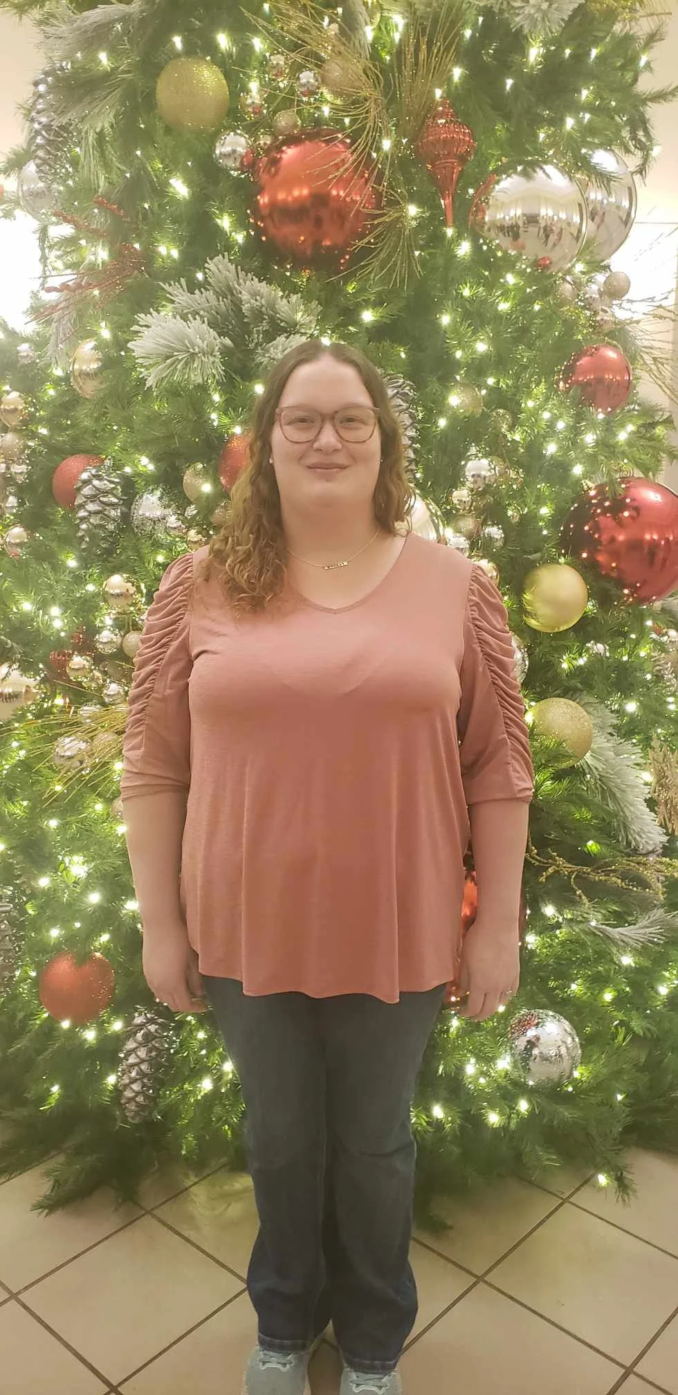 A woman standing in front of a decorated Christmas tree with lights, ornaments, and pinecones.