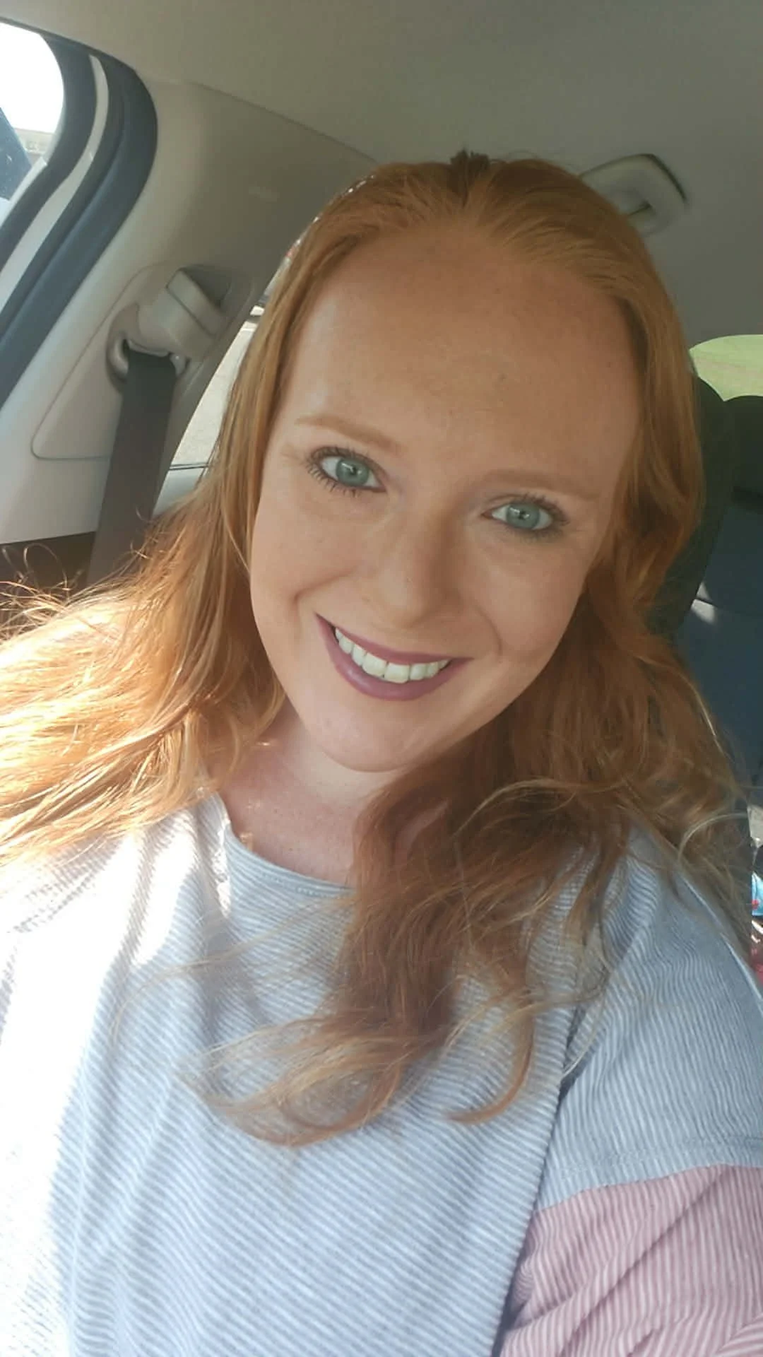 A woman with red hair and blue eyes smiling inside a car.