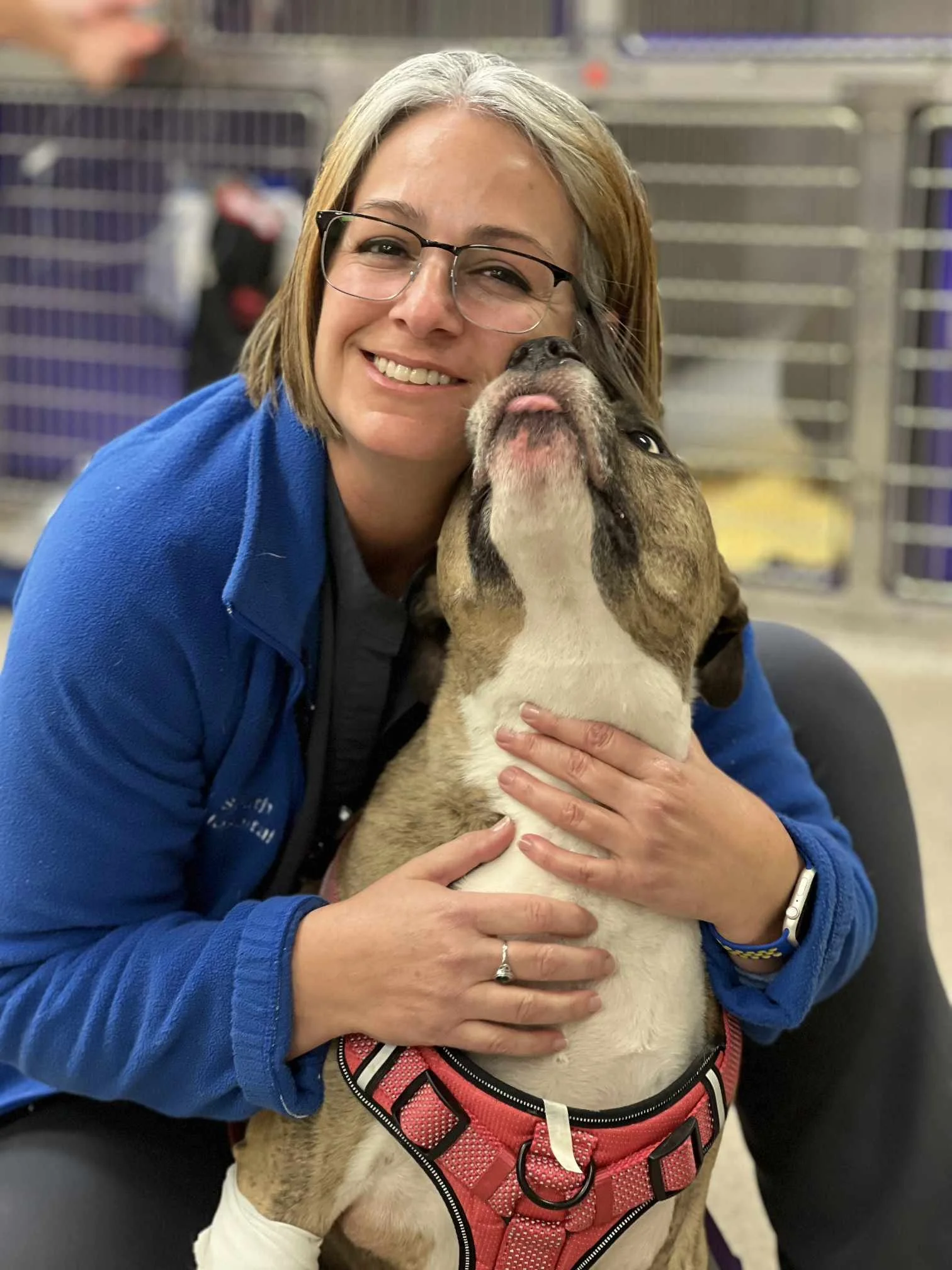 A woman with glasses and a blue jacket hugging a large brindle dog with a pink tongue, in a kennel or shelter setting.