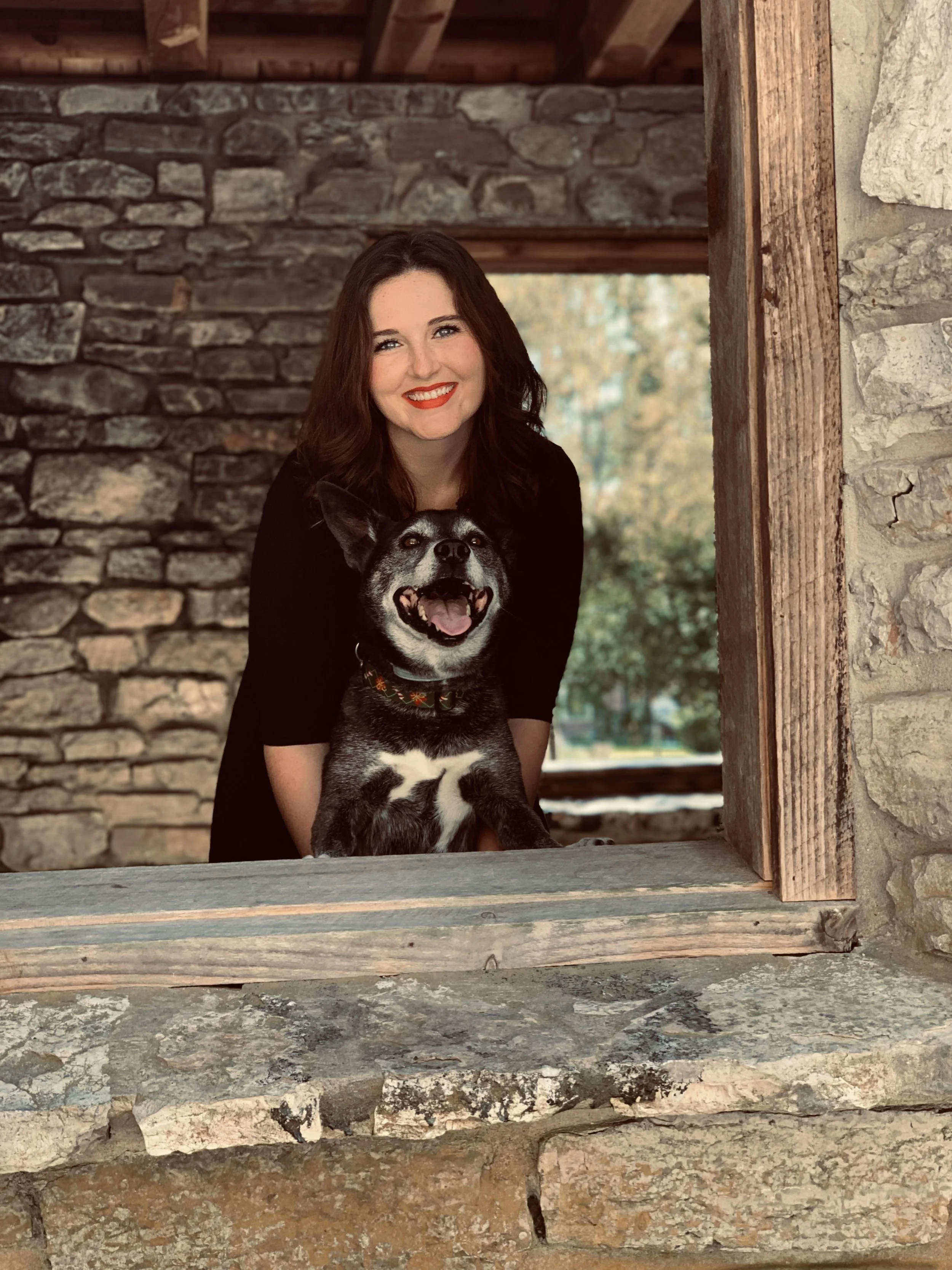 A woman and a black and white dog smiling, posed inside a stone building with a window frame.