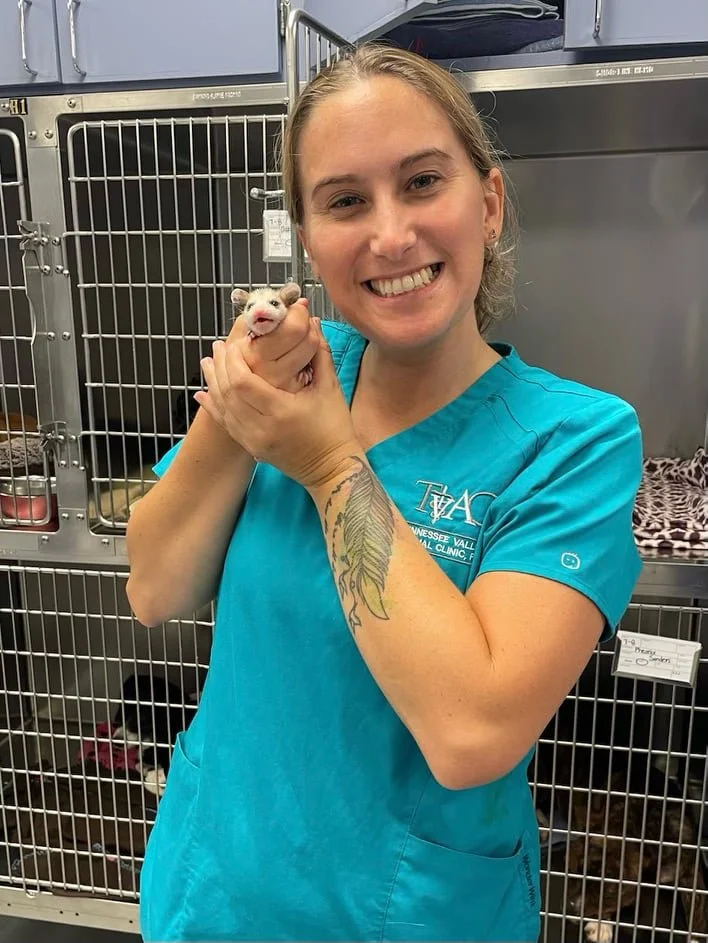 A woman in teal scrubs holding a small ferret in her hands, smiling at the camera in an animal shelter or veterinary clinic, with cages in the background.