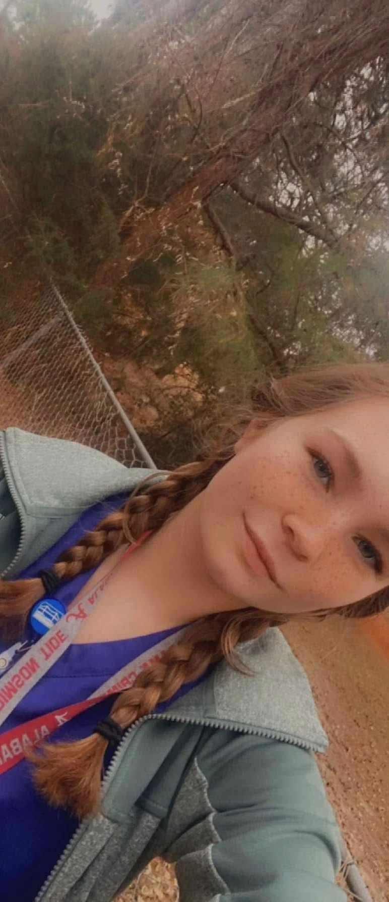 A young woman with braided red hair and freckles taking a selfie outdoors on a trail with trees in the background.