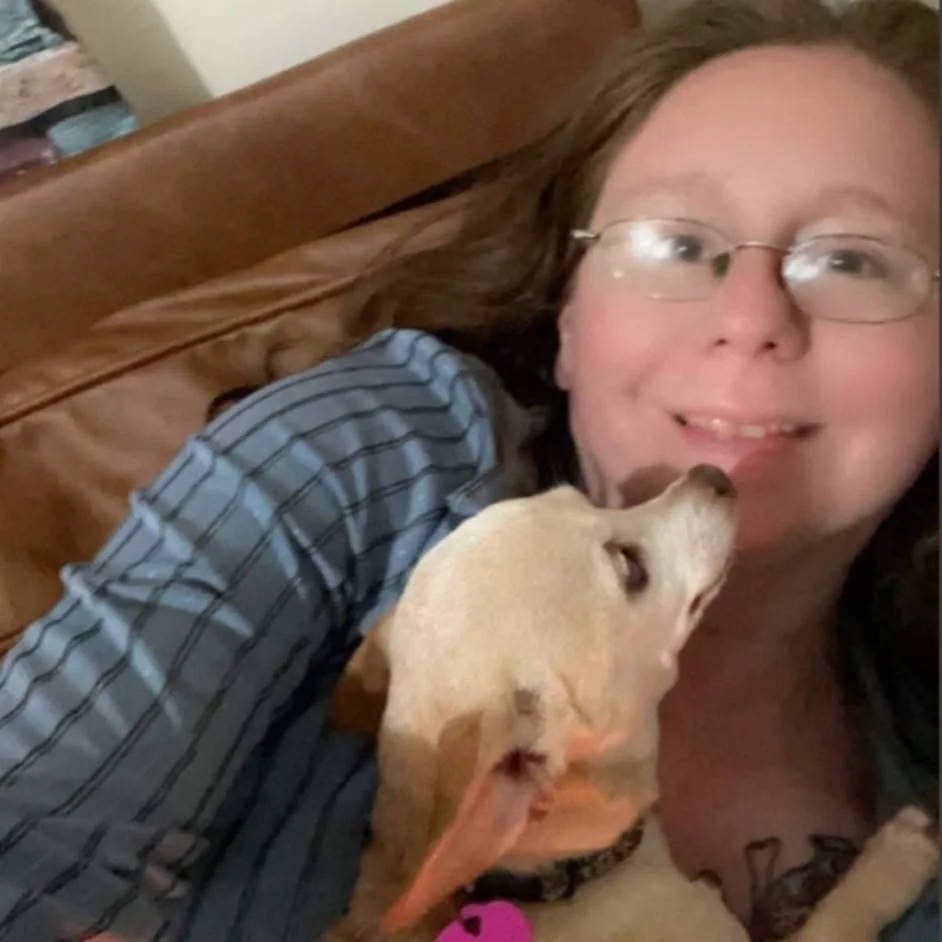 A woman with glasses and long brown hair takes a selfie while lying on a brown leather couch. She is smiling and a tan-colored dog is licking her face.