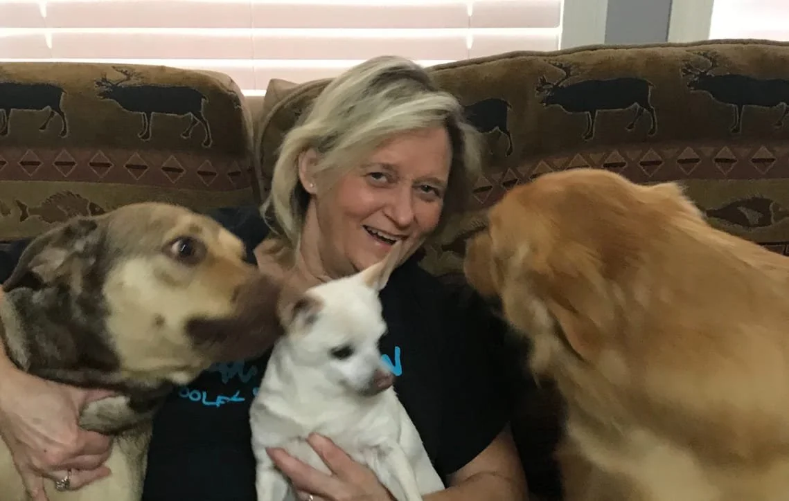 A woman sitting on a couch holding a small white dog, with a large yellow retriever and a large brindle-colored dog close by, in front of a patterned couch with animal motifs.