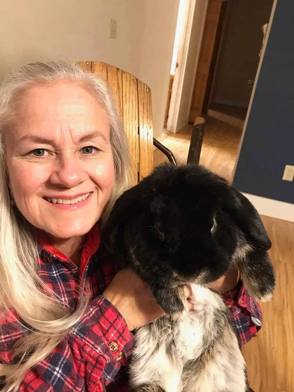 A woman with long blonde hair and a red plaid shirt smiling while holding a black bunny with tan markings in her arms inside a home.