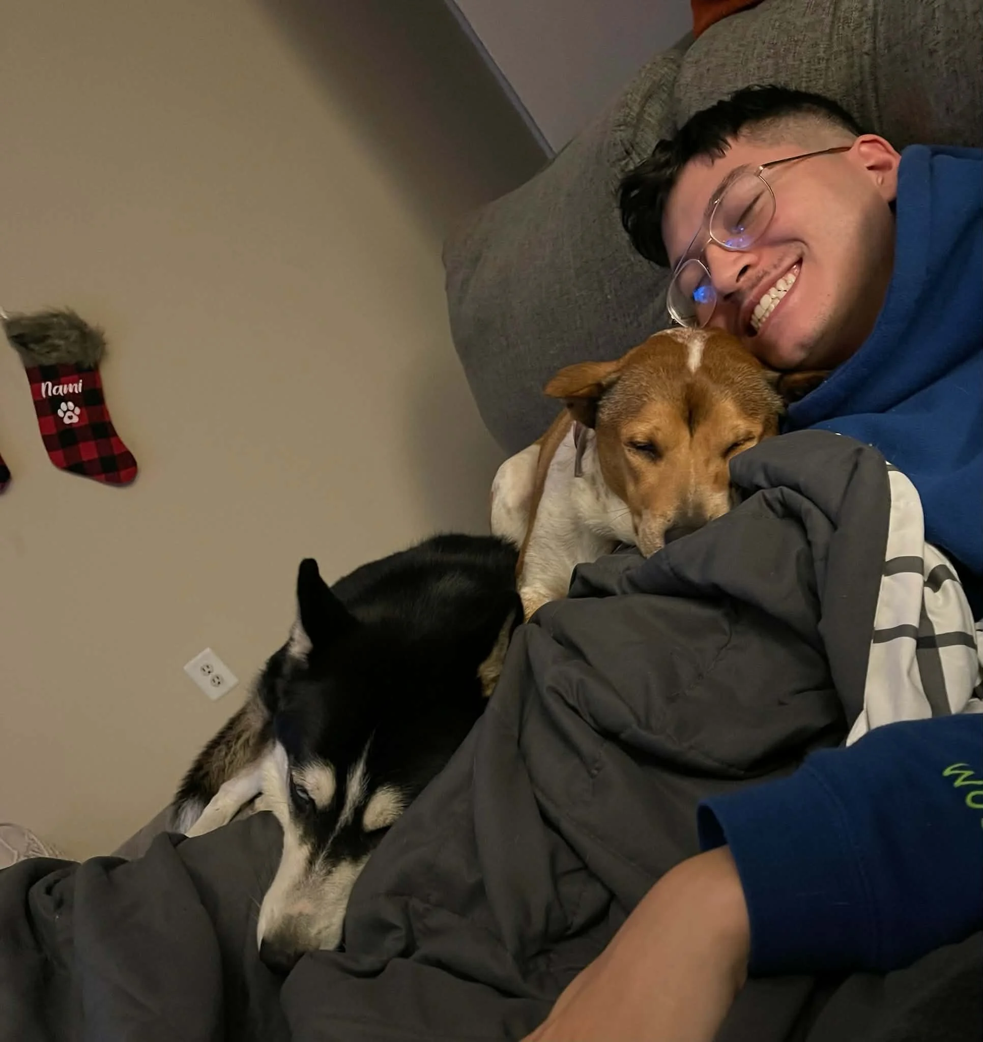 A young man with glasses lying on a couch, smiling, surrounded by two dogs, a Siberian Husky and a mixed breed, all cuddling together.