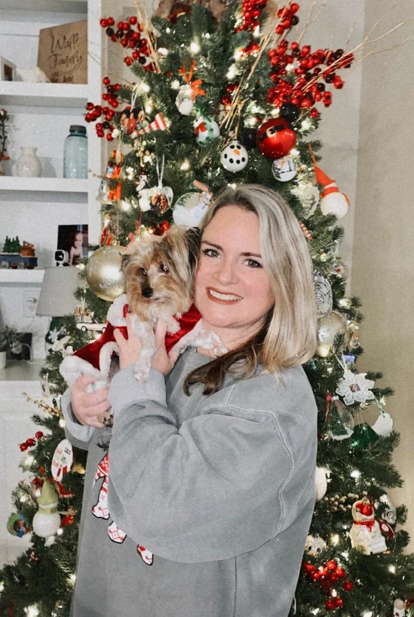 A woman with blonde hair smiling and holding a small dog dressed in a red and white holiday outfit, standing in front of a decorated Christmas tree with red, white, and gold ornaments and lights.