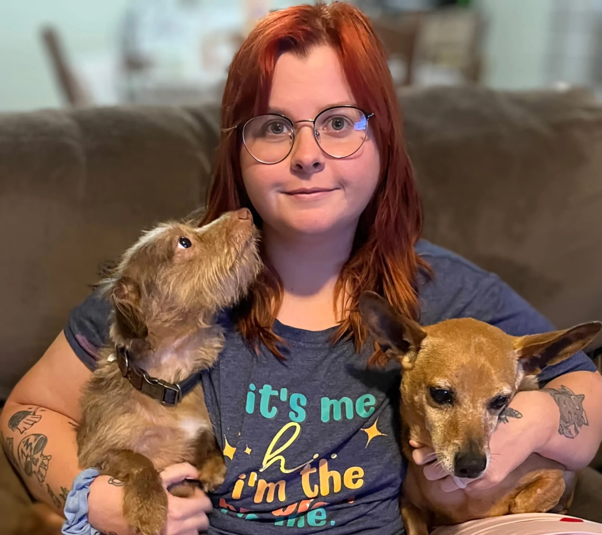 A woman with red hair and glasses holding two small dogs, one in each arm, sitting on a brown couch in a living room.