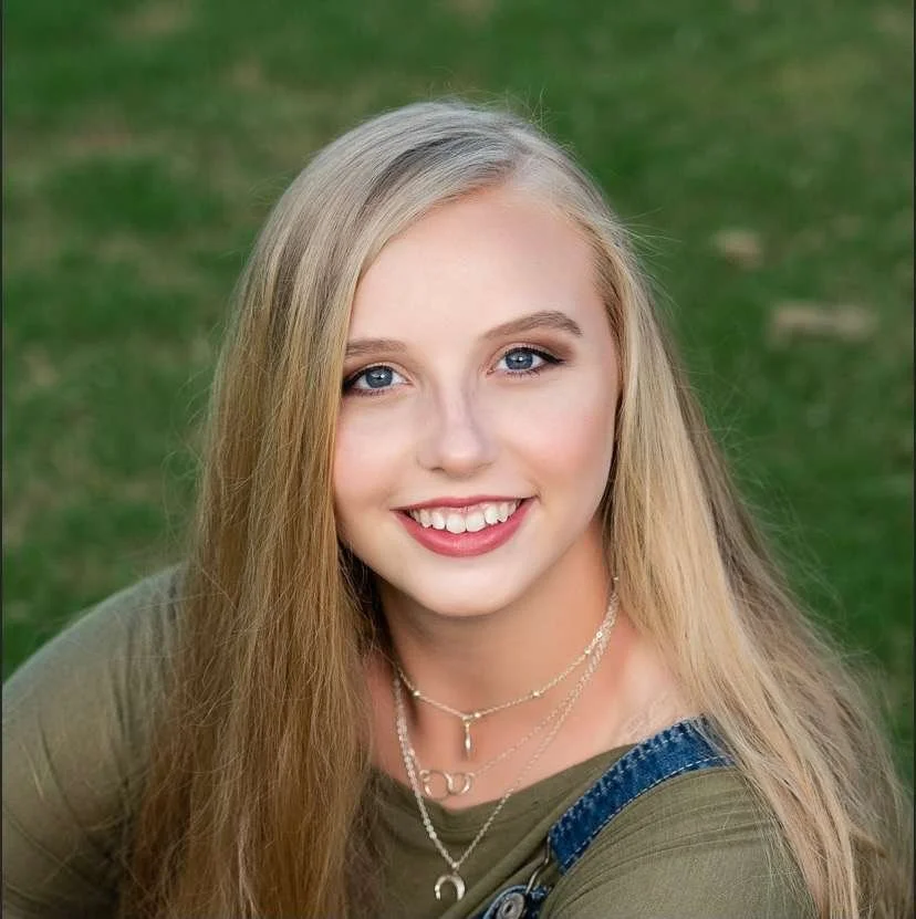 A young woman with long blonde hair smiling outdoors with a grass background.