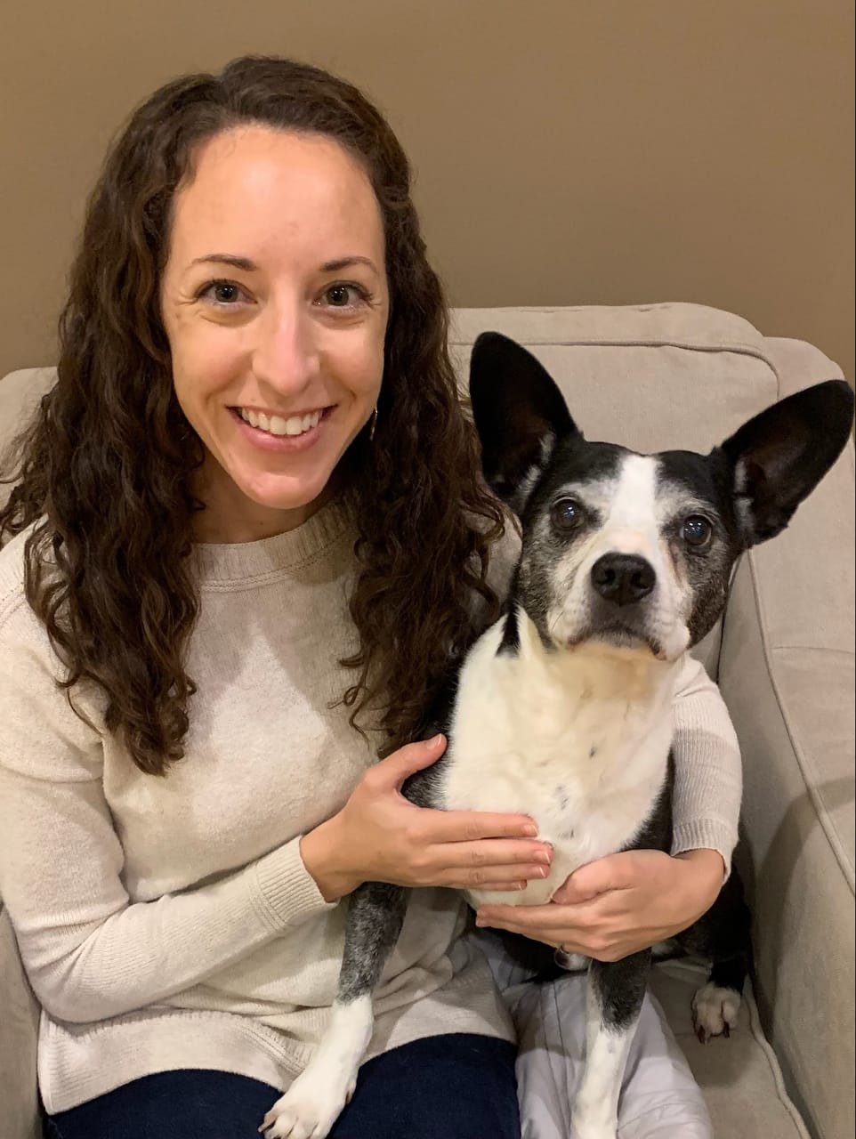 A woman with curly brown hair smiling while holding a black and white dog on her lap, sitting on a beige sofa.