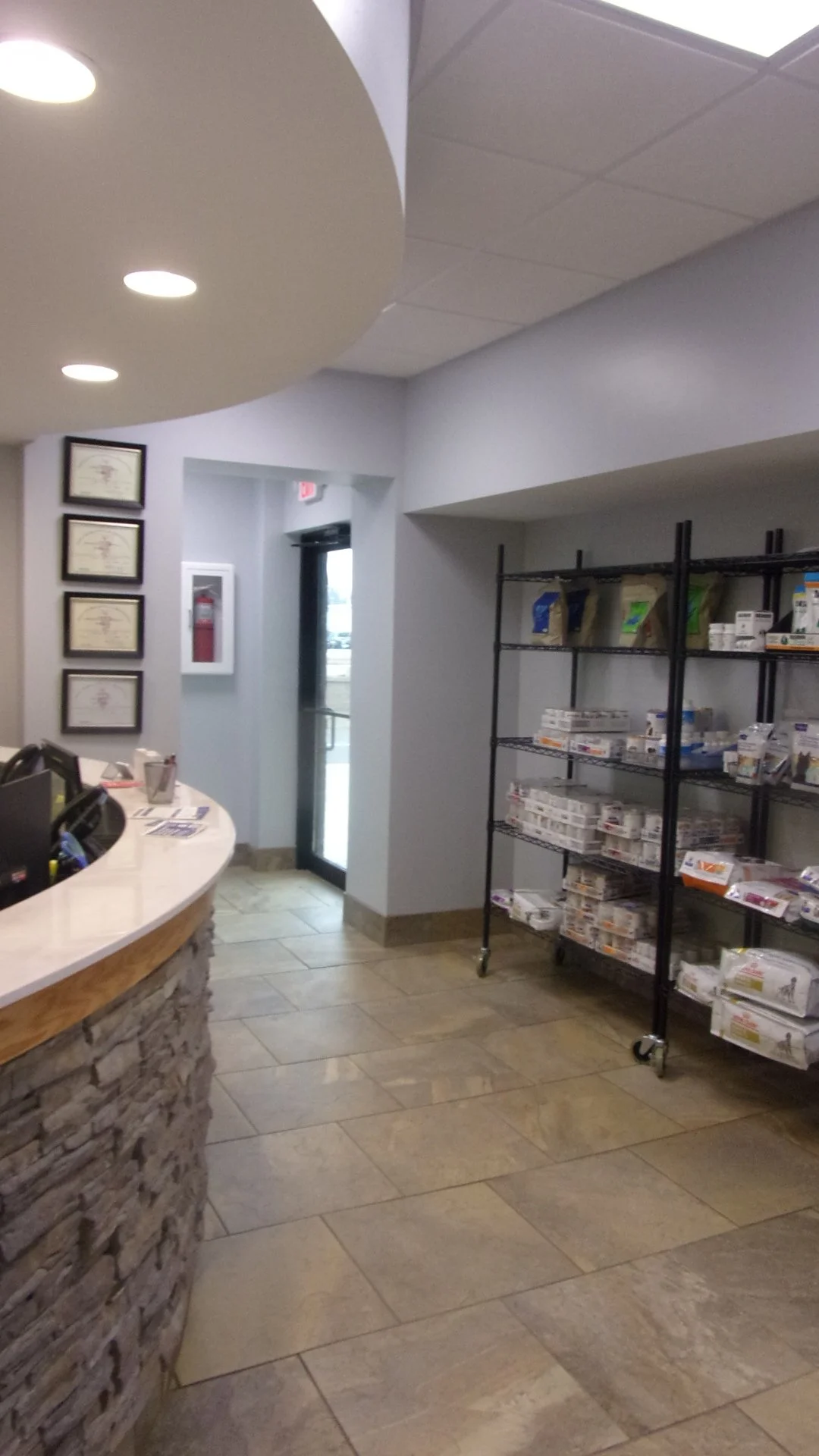 Interior of a medical or dental office reception area with a stone-faced front desk on the left, certificates on the wall, a black metal shelving unit with medical or dental supplies on the right, and an elevator door in the background next to a fire emergency box.