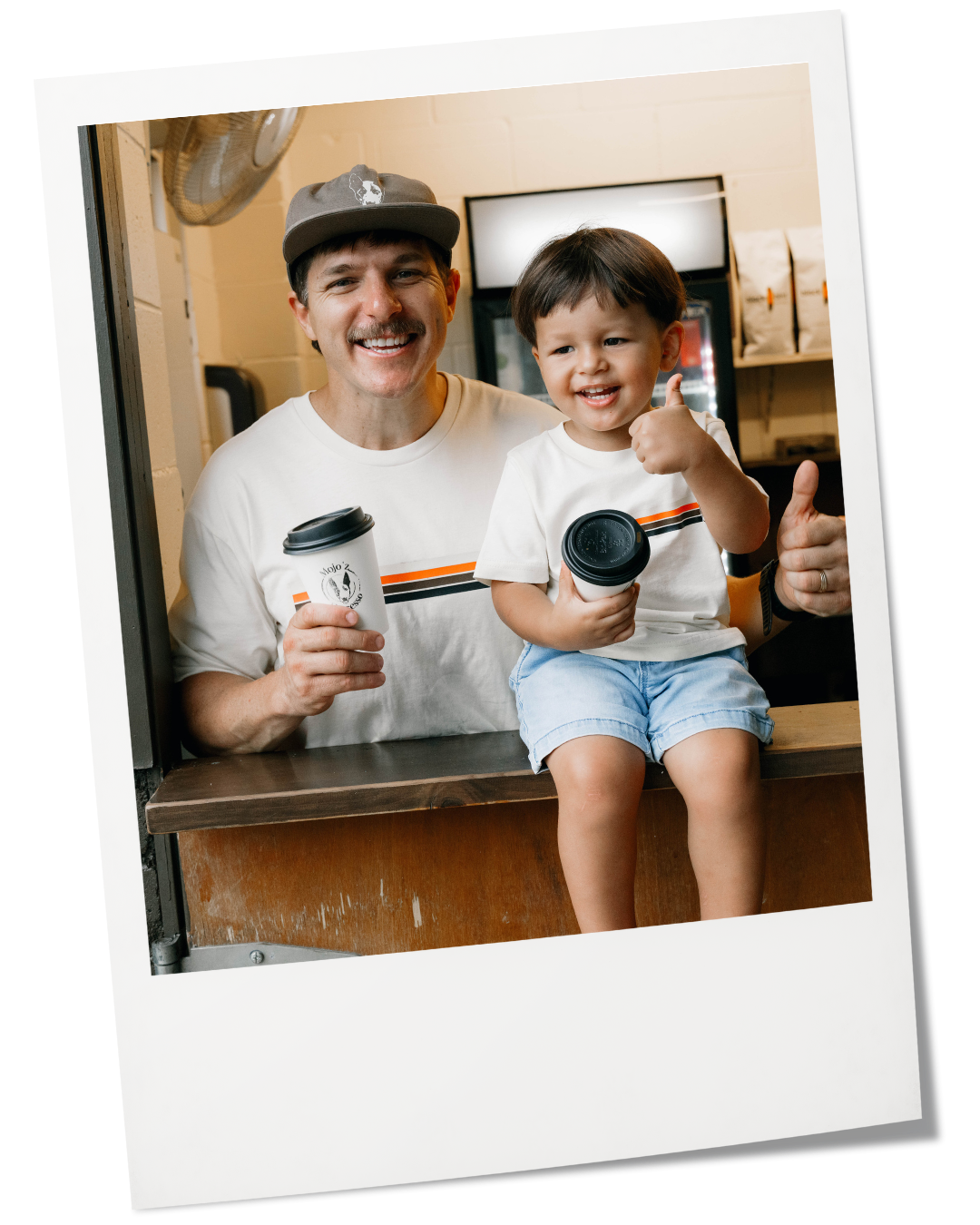 A smiling man and a young boy sitting at a counter holding coffee cups, giving a thumbs-up gesture, in a casual indoor setting.