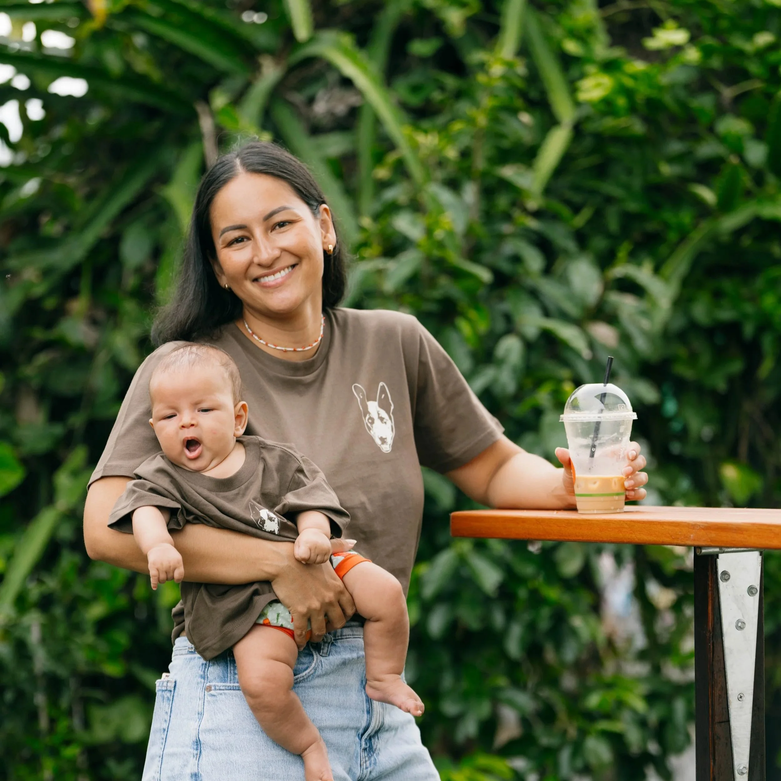 A woman holding a baby in an outdoor setting with green foliage in the background. The woman is smiling and wearing a brown T-shirt with a dog face print. She has a drink on a table next to her.