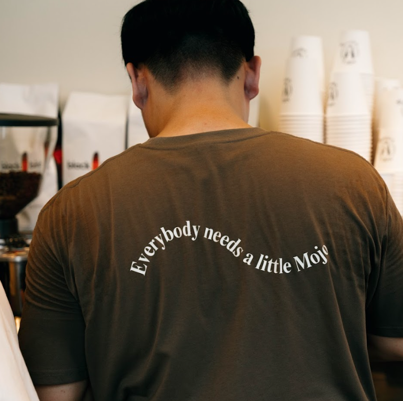 Back of a person wearing a brown T-shirt with white text that reads, 'Everybody needs a little Mojo', near a coffee station with cups and coffee bags.