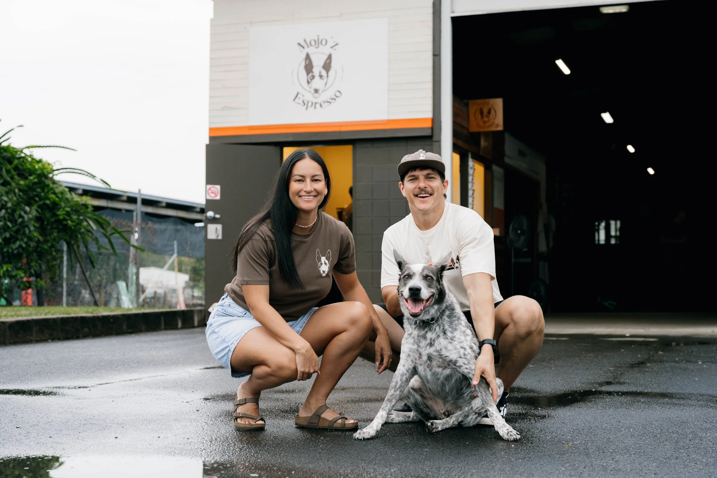 A smiling woman with long black hair, wearing a brown t-shirt and shorts, crouches next to a smiling man with a mustache, wearing a white t-shirt and a cap. They are outdoors on a wet pavement, with a black and white dog sitting between them. Behind them is a building with a sign that says 'Mojo'z Espresso.'