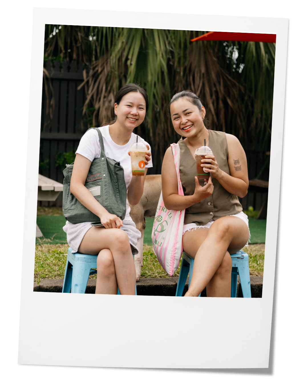 Two women sitting on blue stools outdoors, holding iced coffees, smiling, with a black fence and green plants in the background.