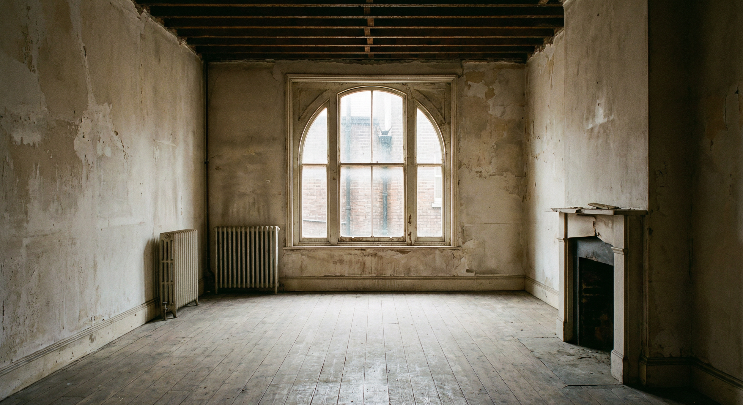 An old, empty room with peeling paint walls, a large arched window, two radiators, and a fireplace with a cracked mantle.