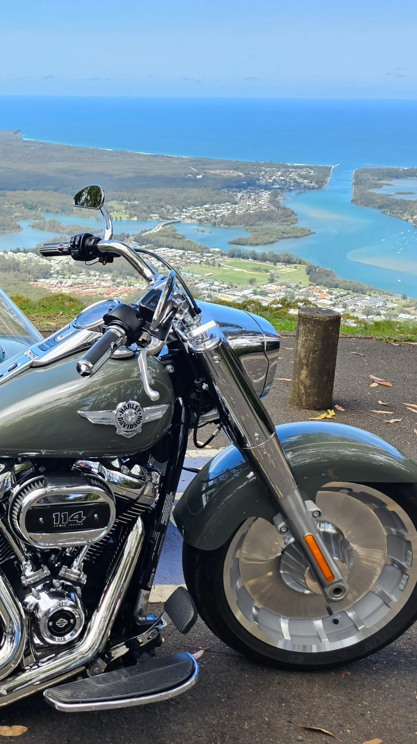 A Harley Davidson motorcycle parked on a scenic overlook with a view of the coastline, water, and a town below