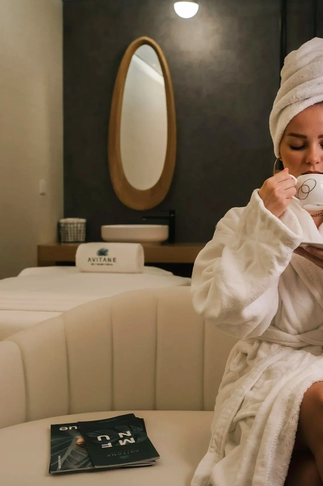 A woman in a white bathrobe and towel wrap drinking from a teacup in a spa room with a bed, mirror, and spa items.
