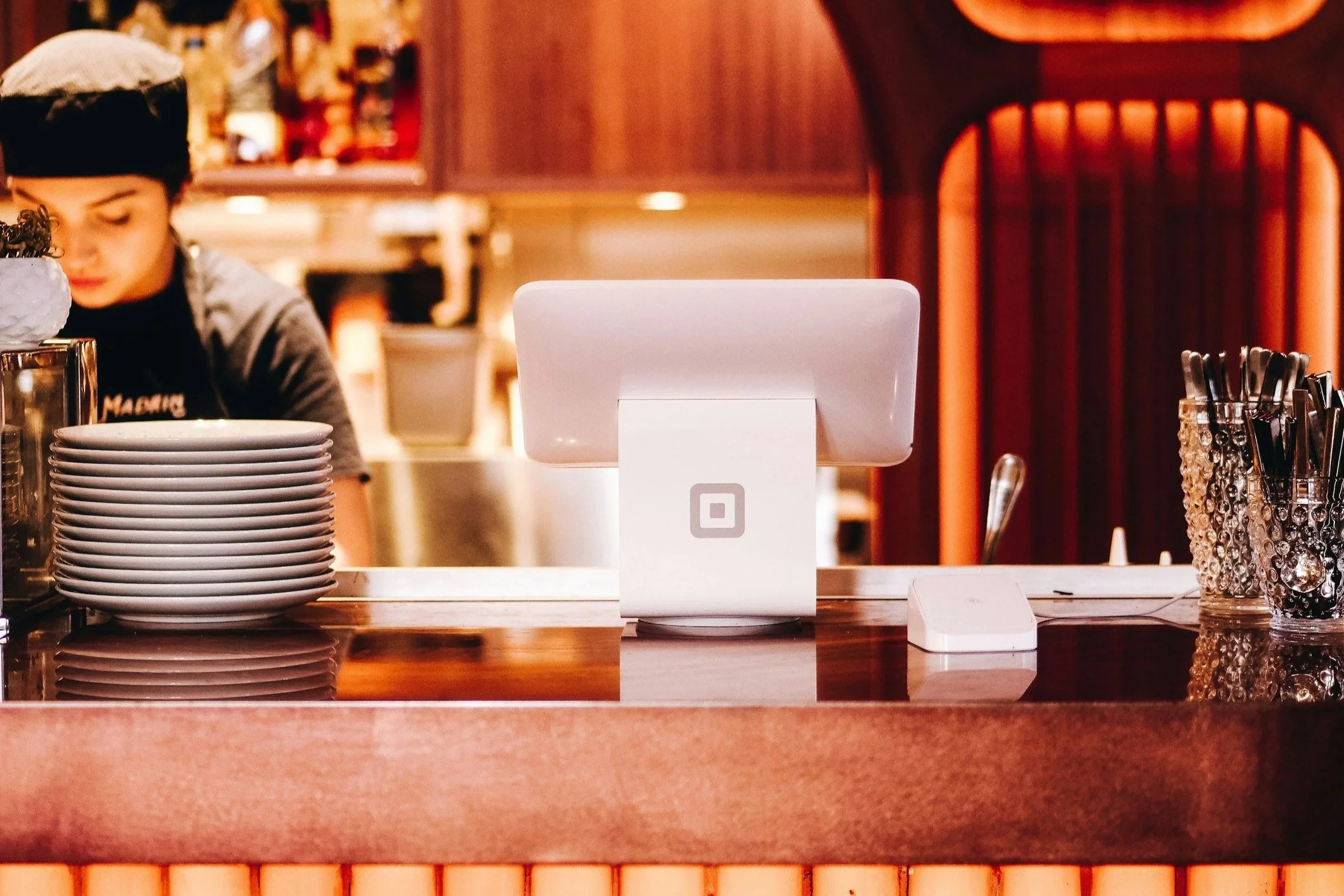A modern point-of-sale terminal on a restaurant counter with stacked plates on the left and a container of utensils on the right, a worker in uniform in the background, and warm wood paneling in the decor.