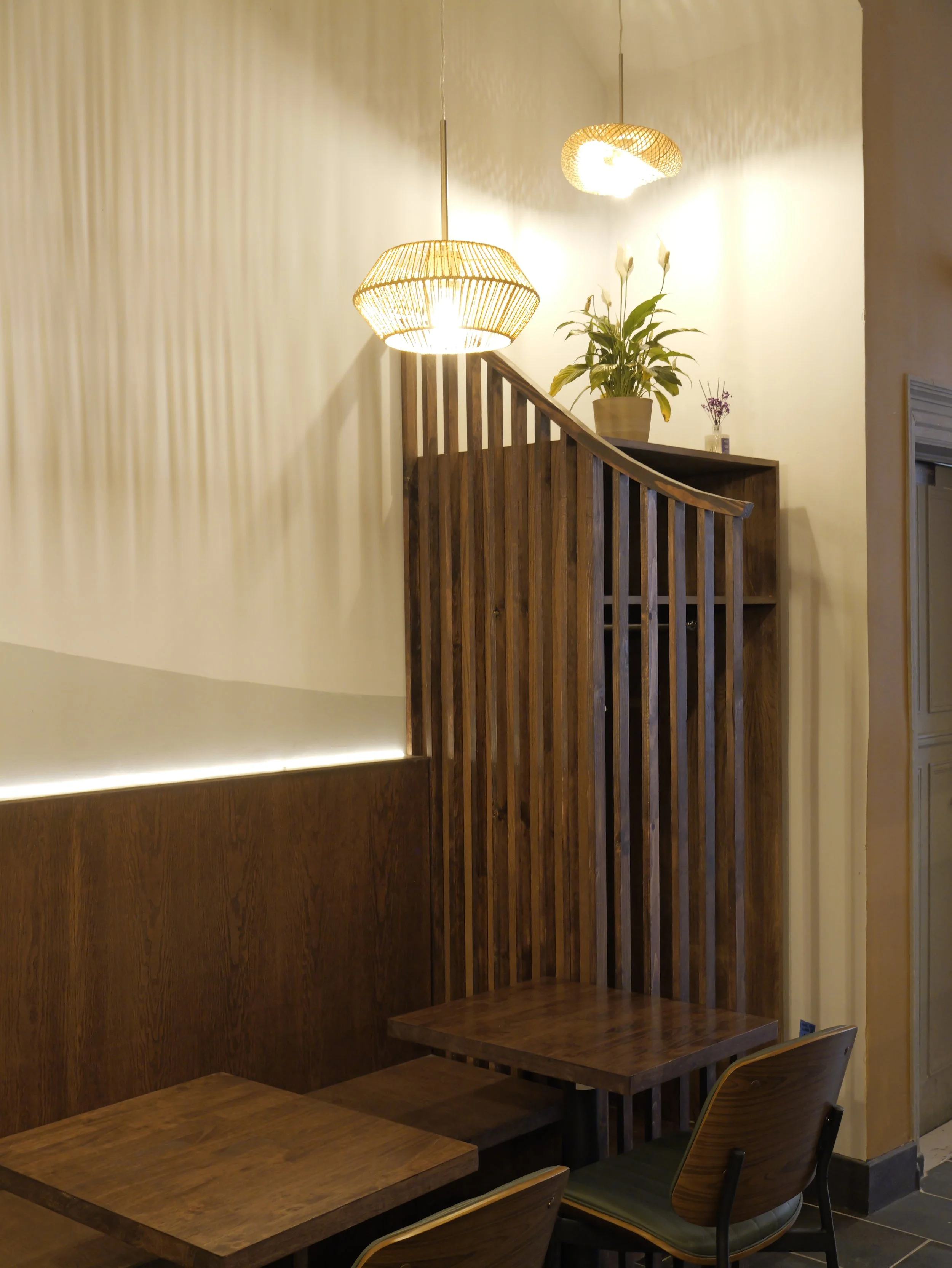 Wooden booth seating with tables and chairs in a cozy restaurant corner, with hanging pendant lights, a potted plant, and decorative flowers on a shelf.