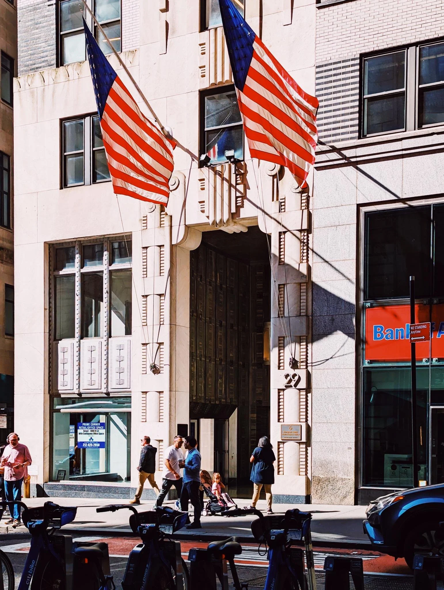 Two American flags hanging on the front of a building in a busy urban street, with people walking and a parked car.