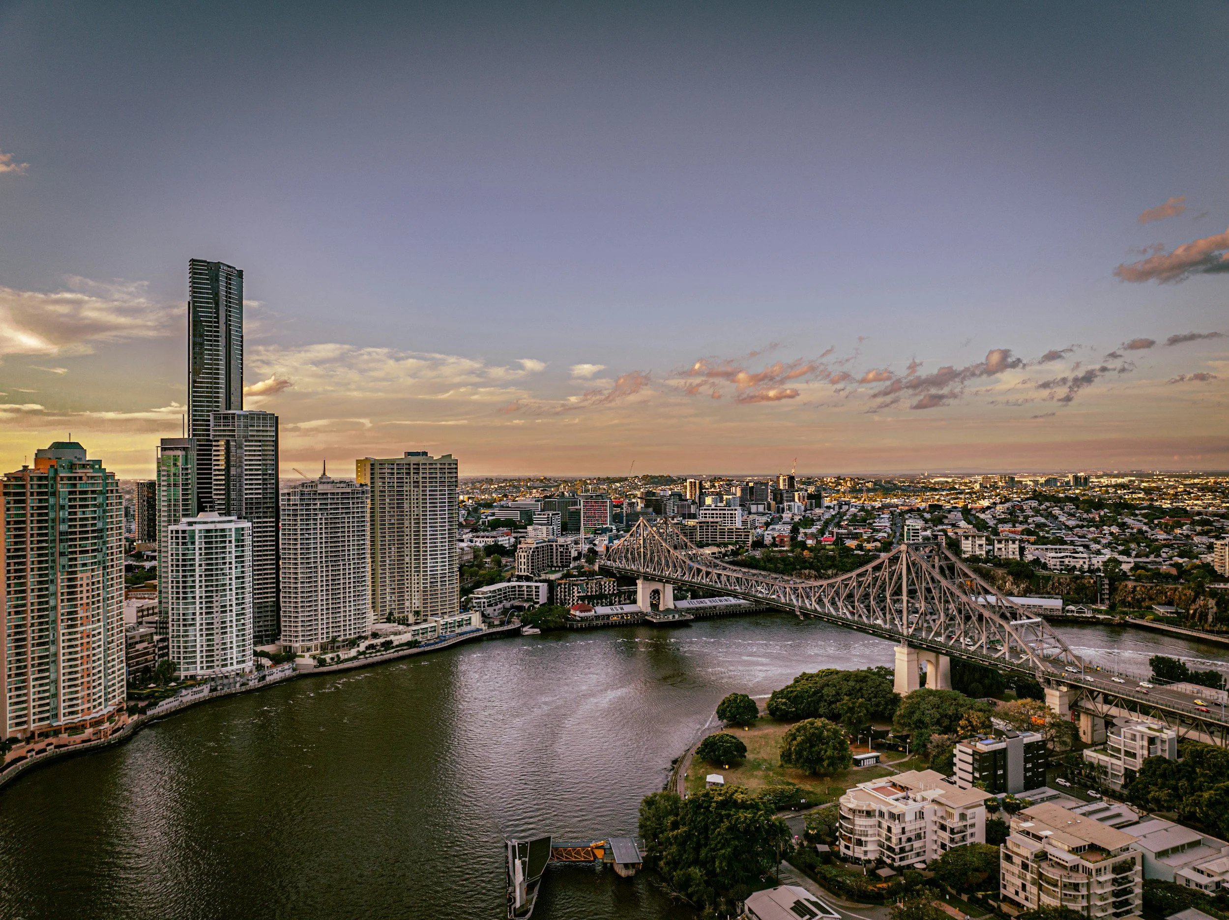 A city skyline at sunset with tall skyscrapers, a river running through the city, and a bridge crossing over the river.