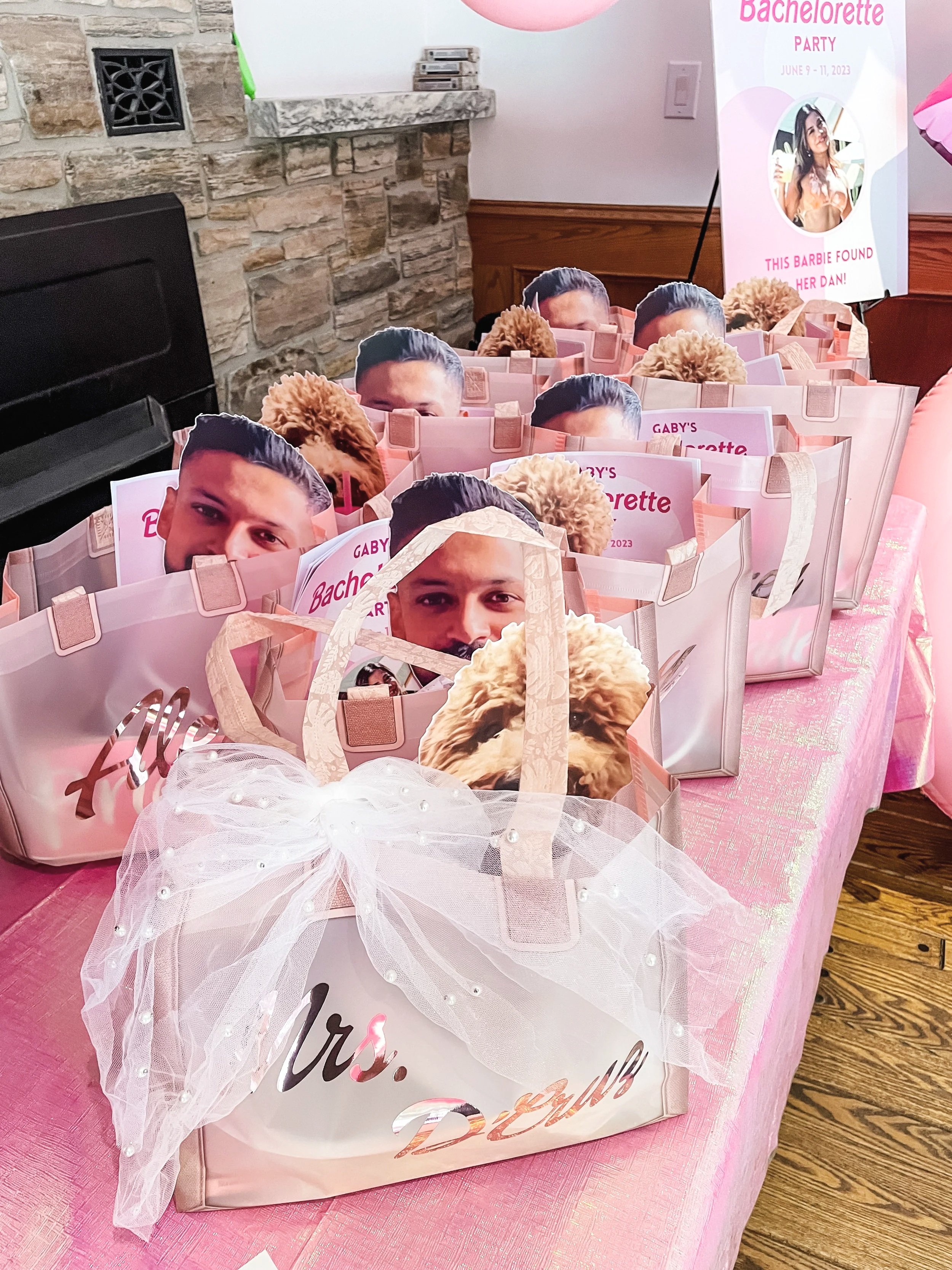 Pink gift bags decorated with photos of a woman and a man, with a dog face sticker, some bags have tulle bows, set on a pink table, with a sign indicating a bachelorette party.