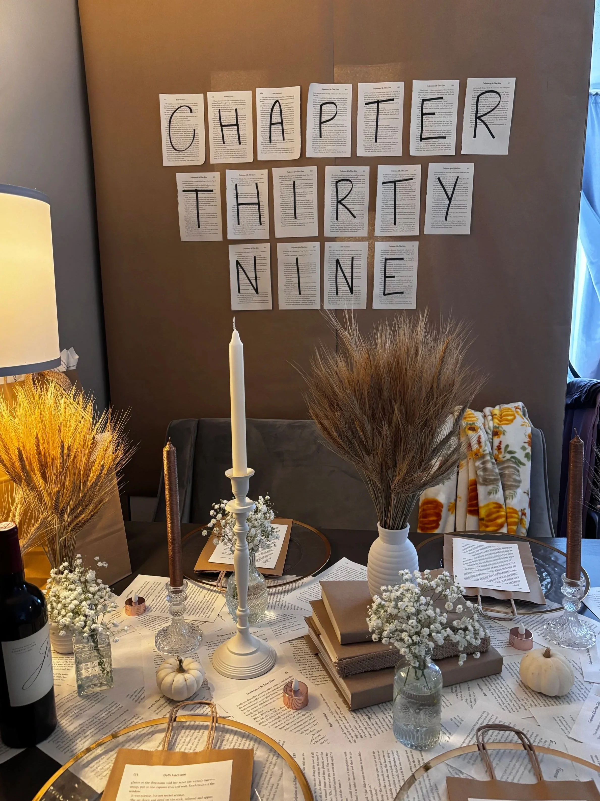 Decorative table setting with books, white pumpkins, and flowers, with a blackout curtain and a wall display of paper pages forming the words 'CHAPTER THIRTY NINE' in black marker for birthday celebration.