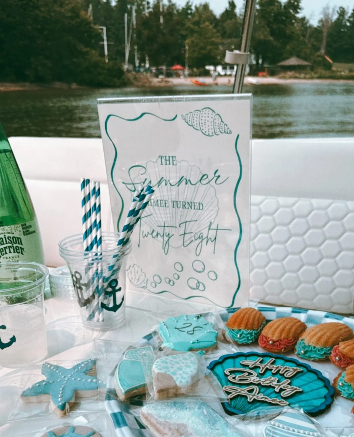 Festive summer-themed party table with cookies, cupcakes, striped straws, and decorative sign reading 'The summer I came turned twenty-eight' near a lake with trees and outdoor scenery in the background.