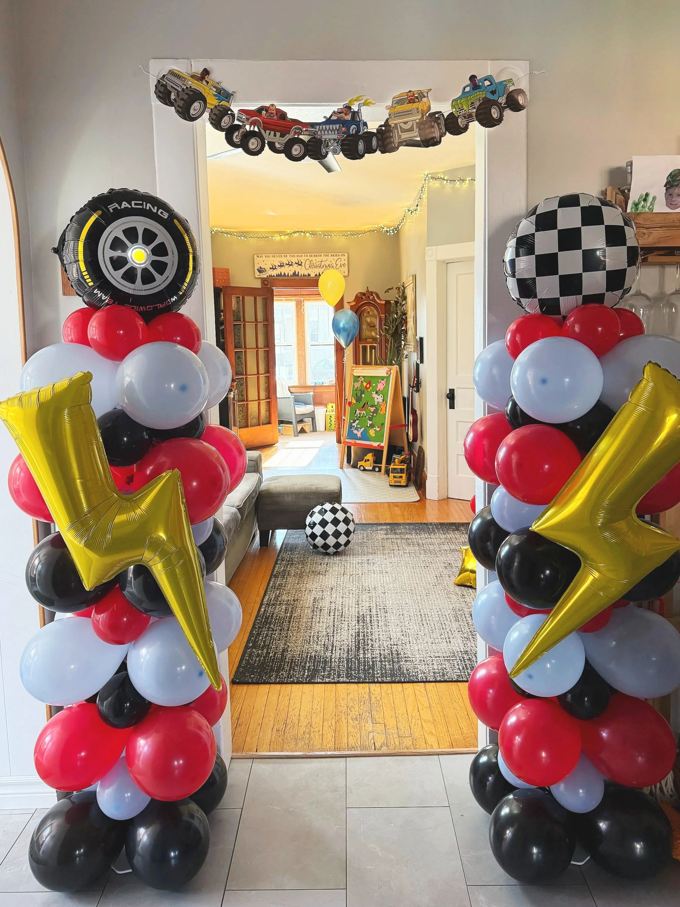 Decorated doorway with racing-themed balloon arch at a child's birthday party, featuring checkered flags, cars, and stars, leading into a living room with balloons and toys.