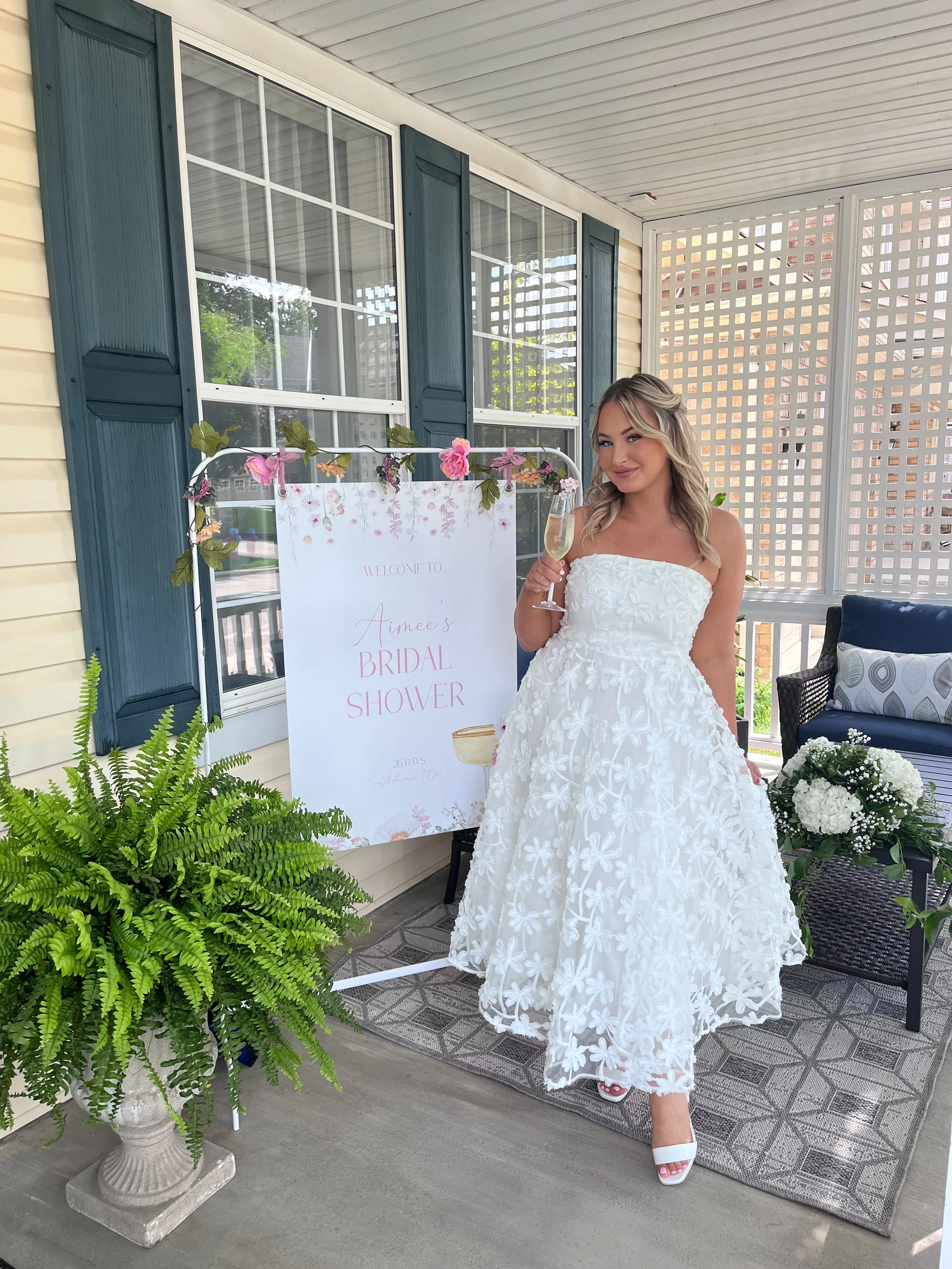 A woman in a white bridal dress holding a glass of champagne at a bridal shower, standing on a patterned rug on a porch with a welcome sign, plants, and outdoor furniture.