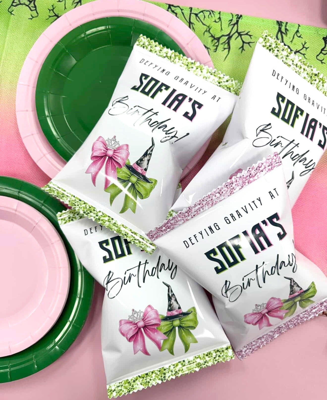 Party favor bags with 'Defying Gravity at Sofia's Birthday' printed on them, decorated with witch hats, pink and green bows, and tiaras, along with pink and green paper plates.