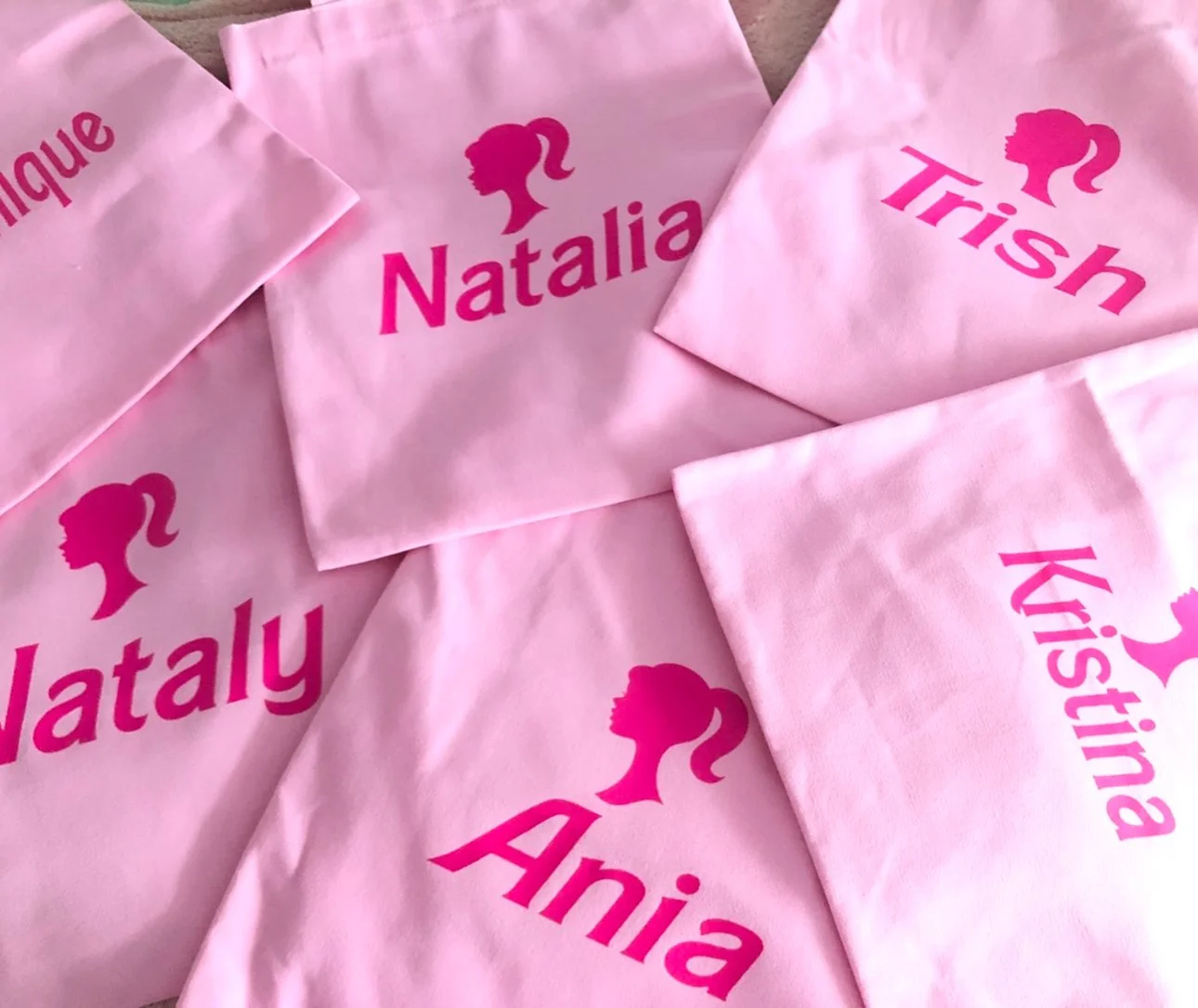Multiple pink cloth gift bags with a silhouette of a barbie girl's profile and names written in bold pink letters, including "Natalia," "Trish," "Kistina," and "Analia."