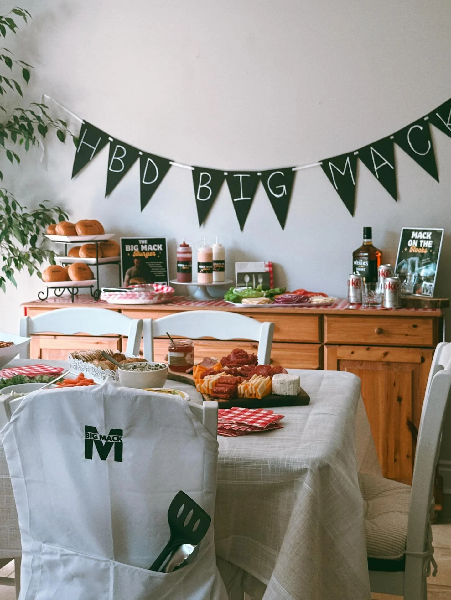 Decorative table set for a casual gathering with a hot dog theme, featuring a banner that reads 'HBD BIG MAC,' food items including cheese, meats, and condiments, and beverages on a wooden sideboard.
