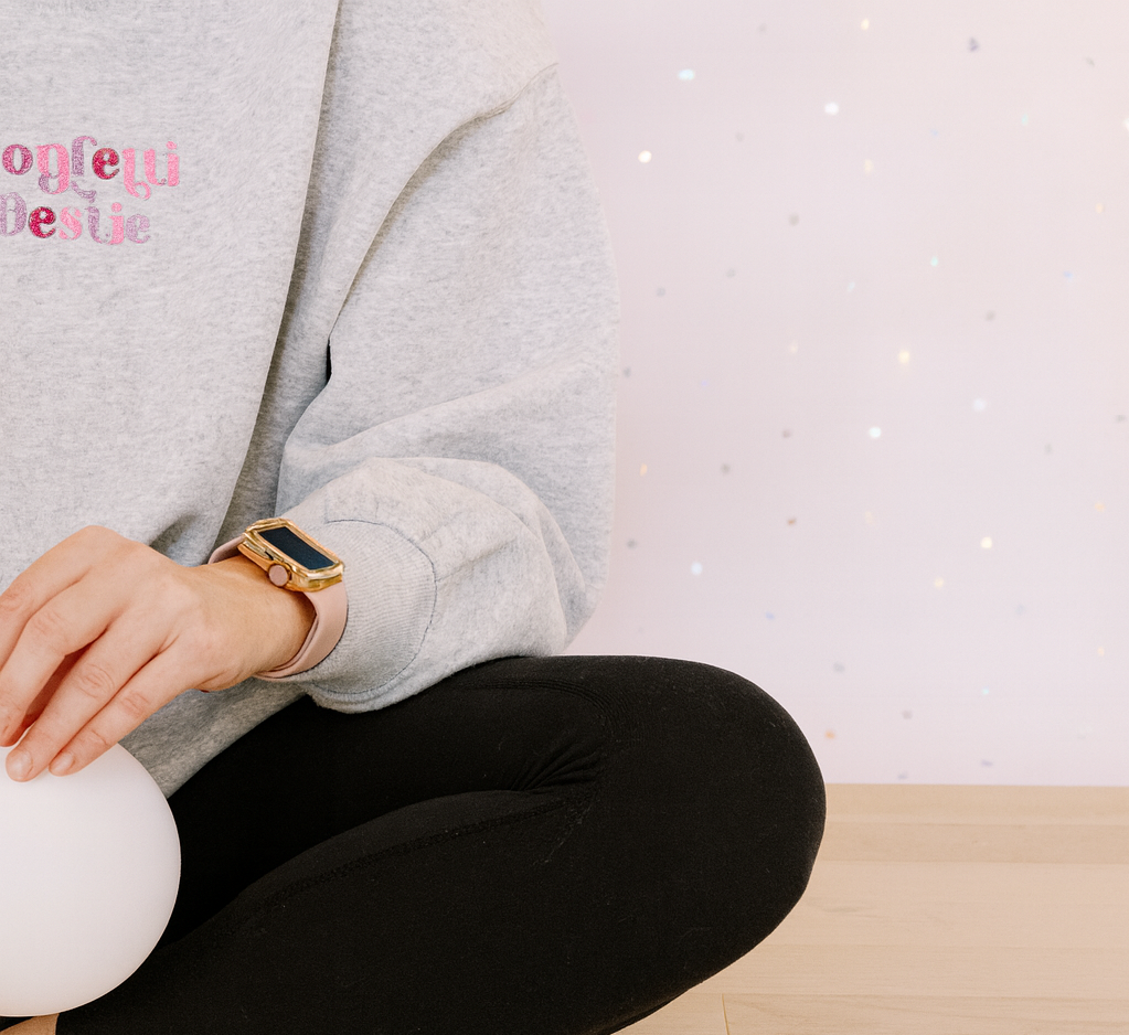 Person sitting cross-legged on wooden floor wearing a gray sweatshirt with pink lettering, black pants, and a smartwatch, holding a white ceramic object, with a light-colored, speckled wall in the background.