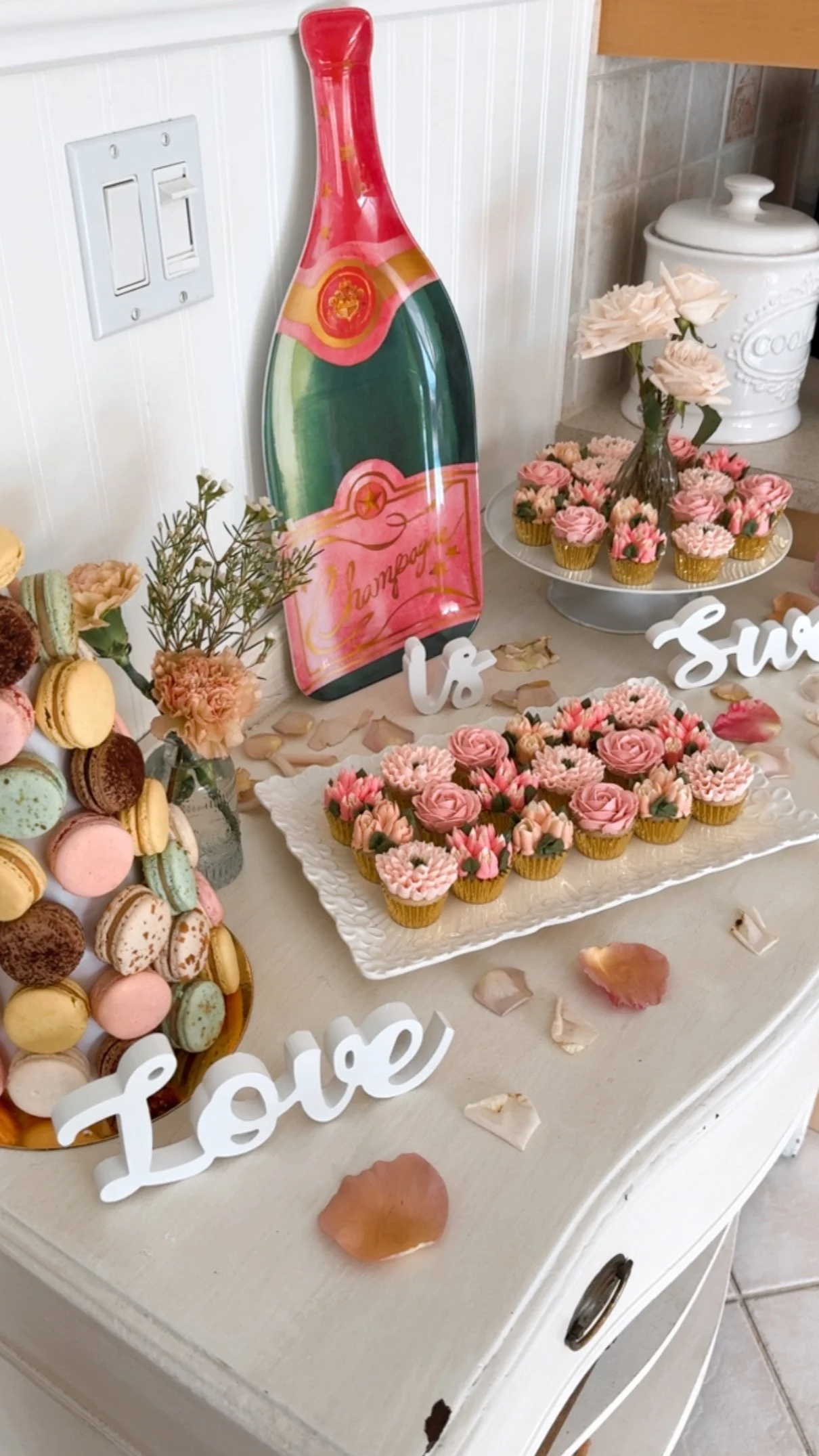 Table display of pink and cream cupcakes, macarons, a wine bottle decorative sign, white and pink floral arrangements, and love-themed decorations with rose petals scattered on the surface.