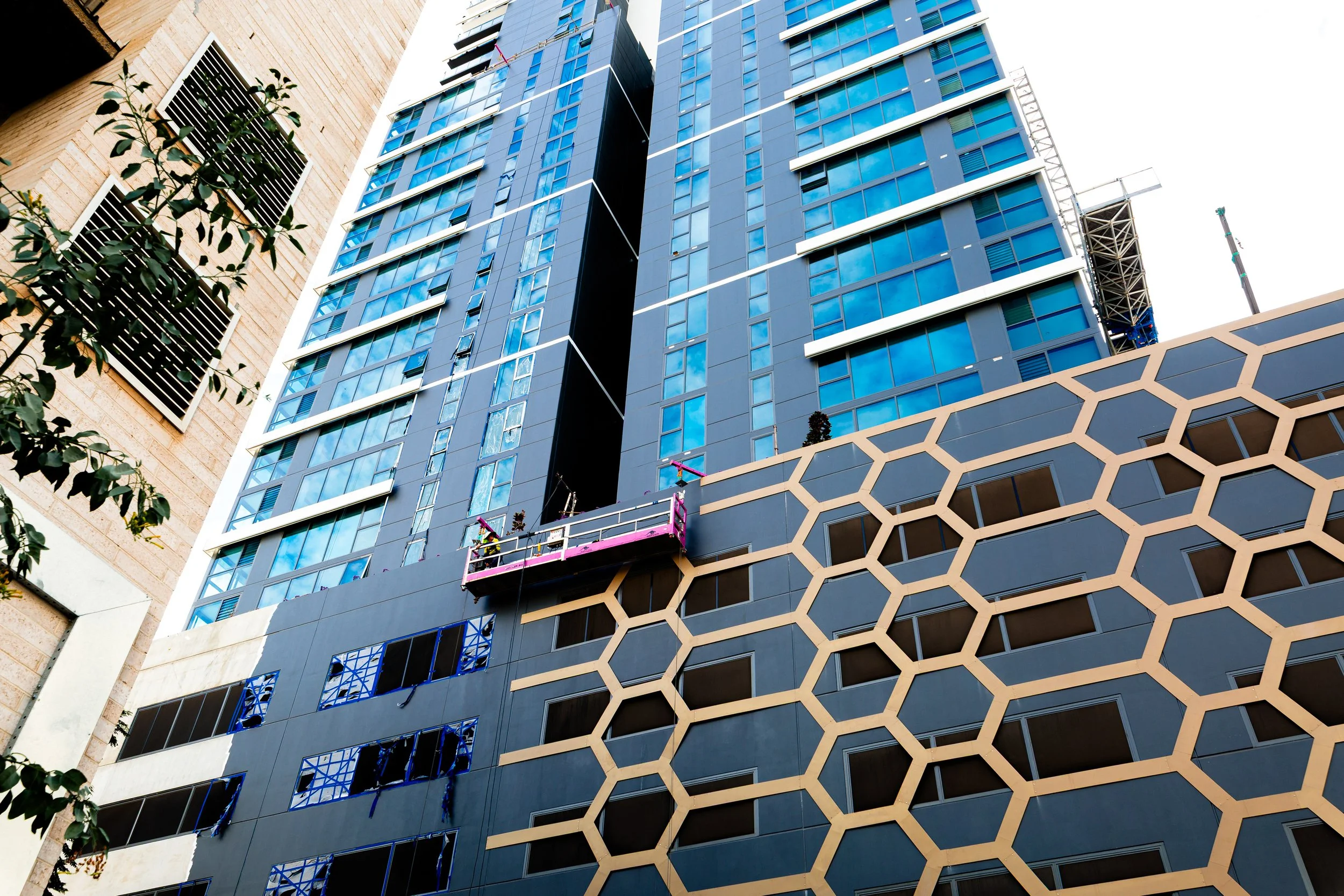 Construction workers on a suspended platform working on the exterior of a modern, high-rise building with glass windows and geometric design panels. AT238 building in Perth CBD. ANCR
