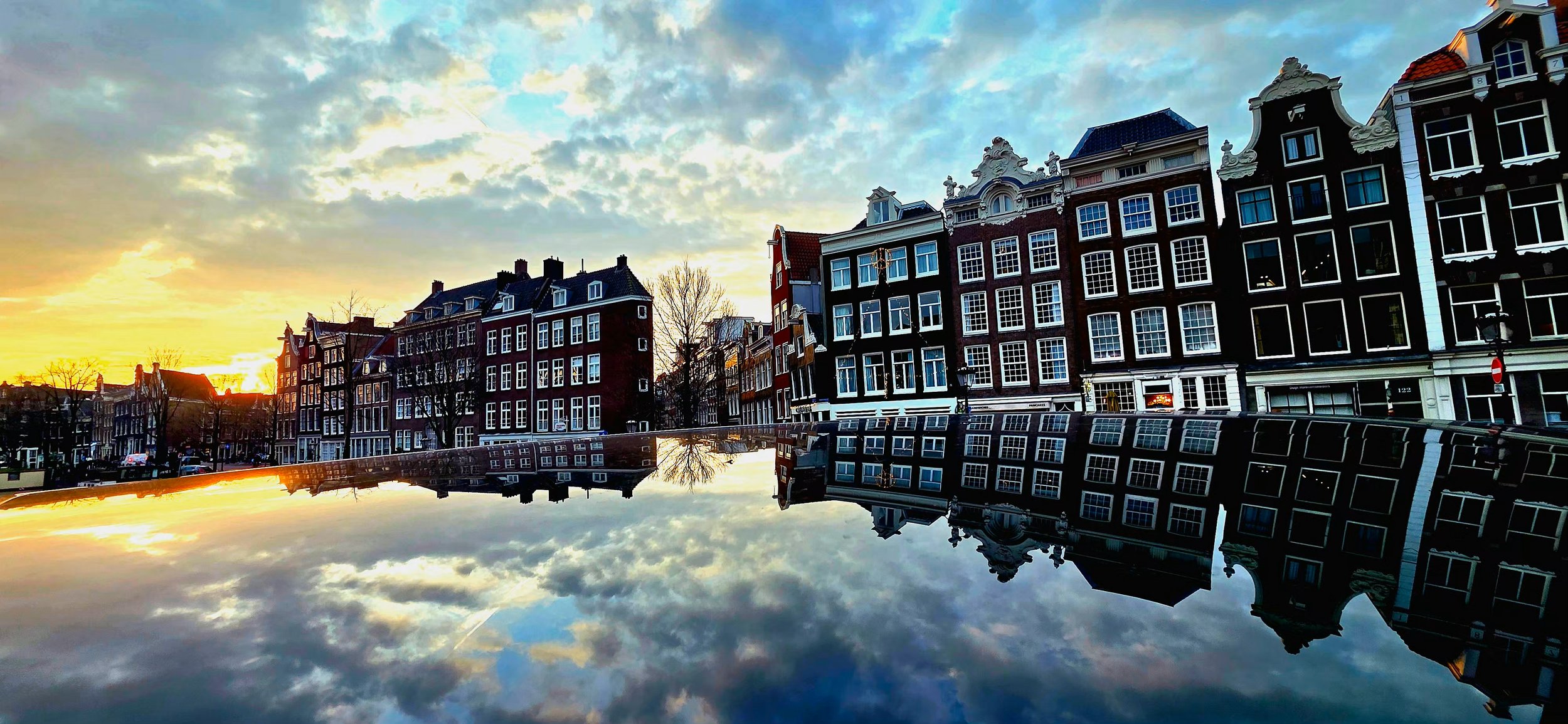 A cityscape at sunset featuring traditional European-style buildings with large windows, reflected in a body of water, with a partly cloudy sky.