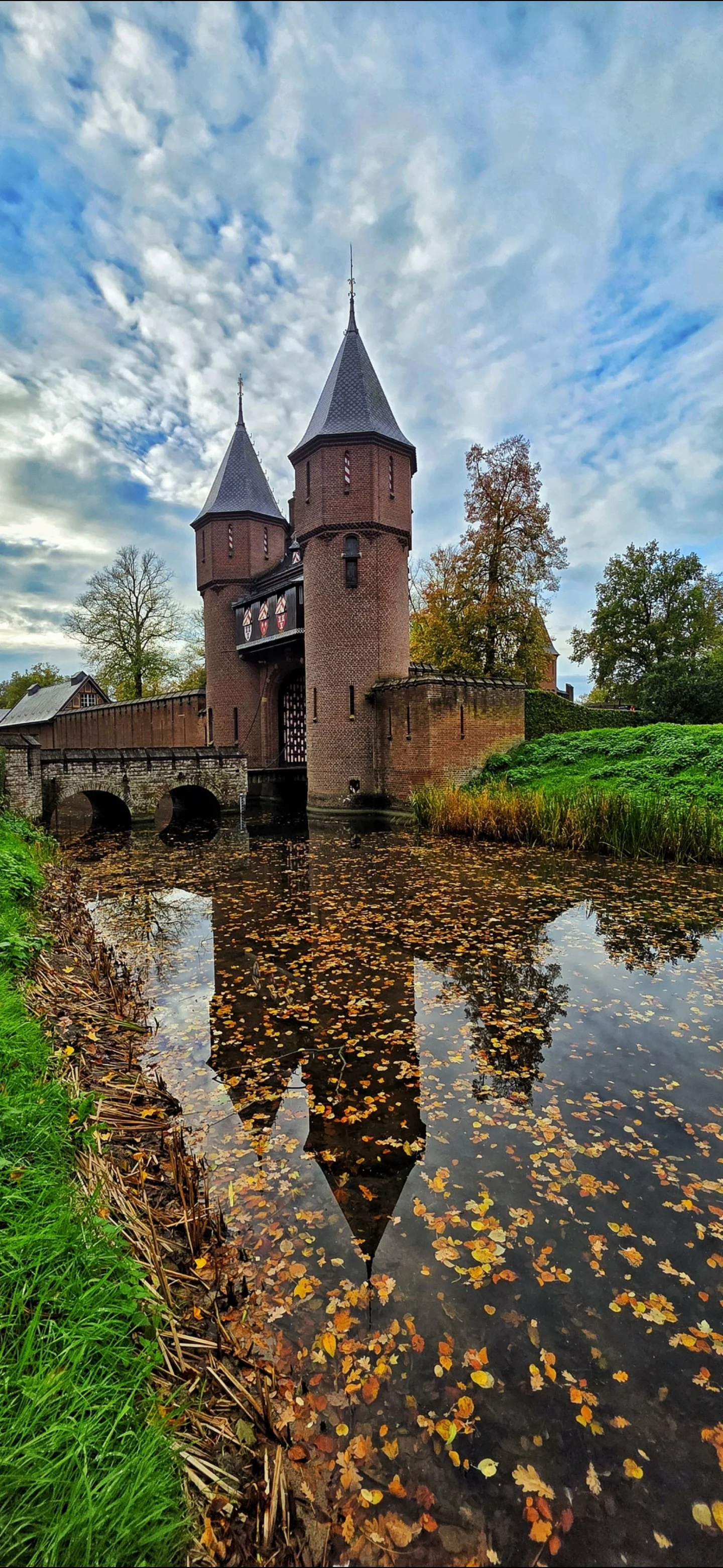 A medieval stone castle with two tall towers featuring conical roofs, set beside a reflective water moat filled with fallen autumn leaves. The sky is partly cloudy with patches of blue, and there are trees with autumn foliage around the castle.