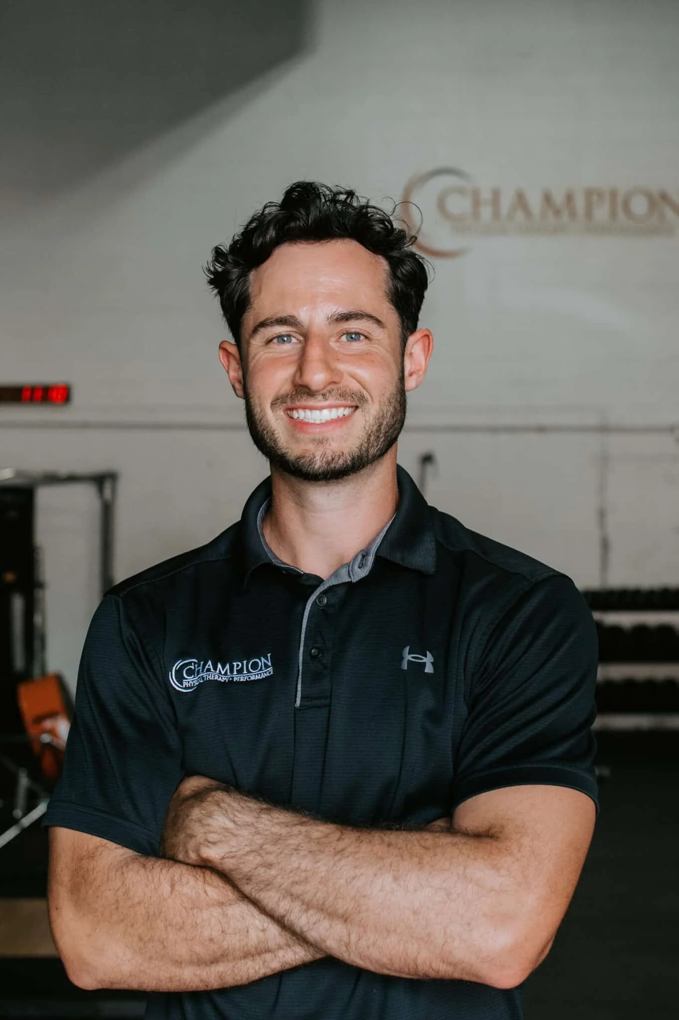 A smiling man with dark hair and beard standing with arms crossed in a gym, wearing a black Under Armour shirt with 'Champion' logo, with gym equipment in the background.
