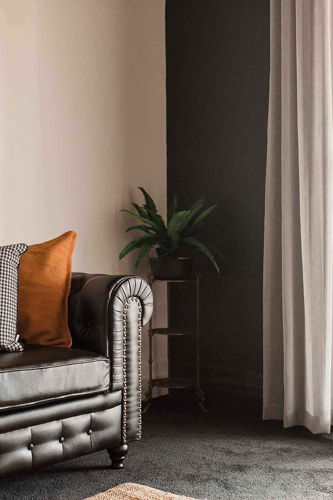 Living room corner with a black leather chesterfield sofa, orange and houndstooth pillows, and a potted green plant on a black metal plant stand next to a window with white curtains.