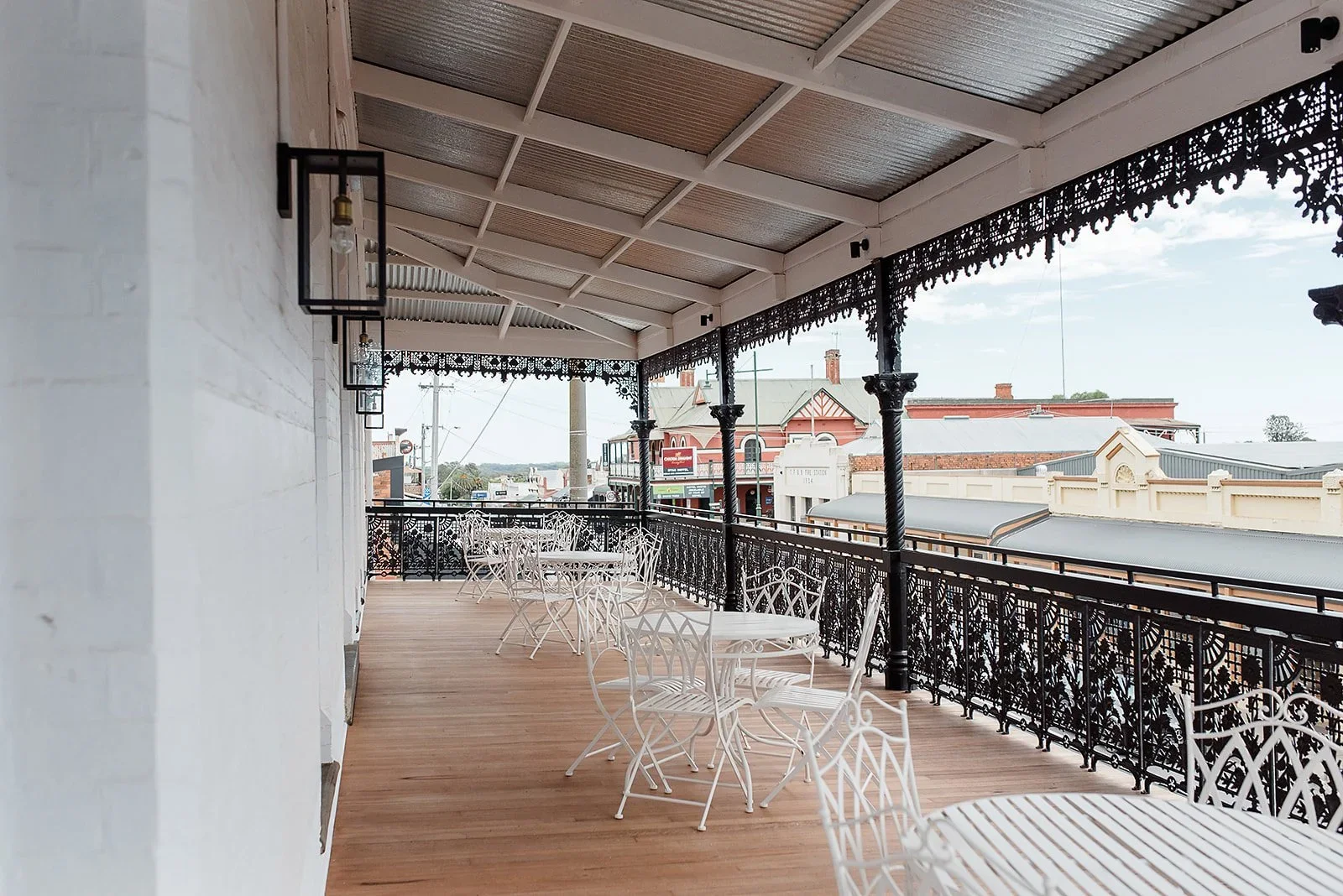 Empty outdoor balcony with white metal chairs and tables, black decorative iron railing and trim, hanging black metal lanterns, on a wooden floor, overlooking a main street with historic buildings, under a sloped roof with white beams and a corrugate