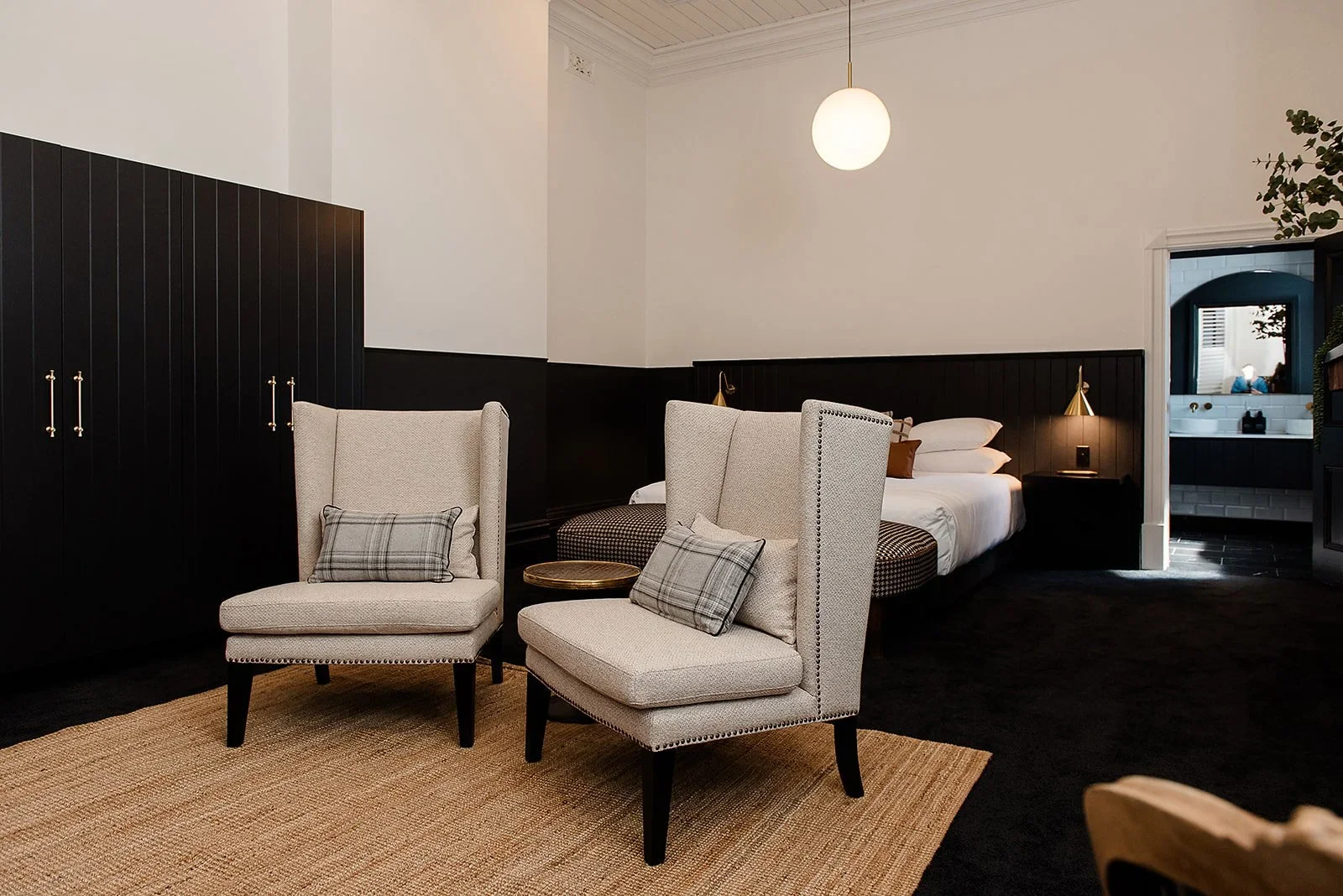 A bedroom with a bed, two beige wingback chairs with plaid pillows, a black wardrobe, and a doorway leading to a bathroom with a white sink and mirror.