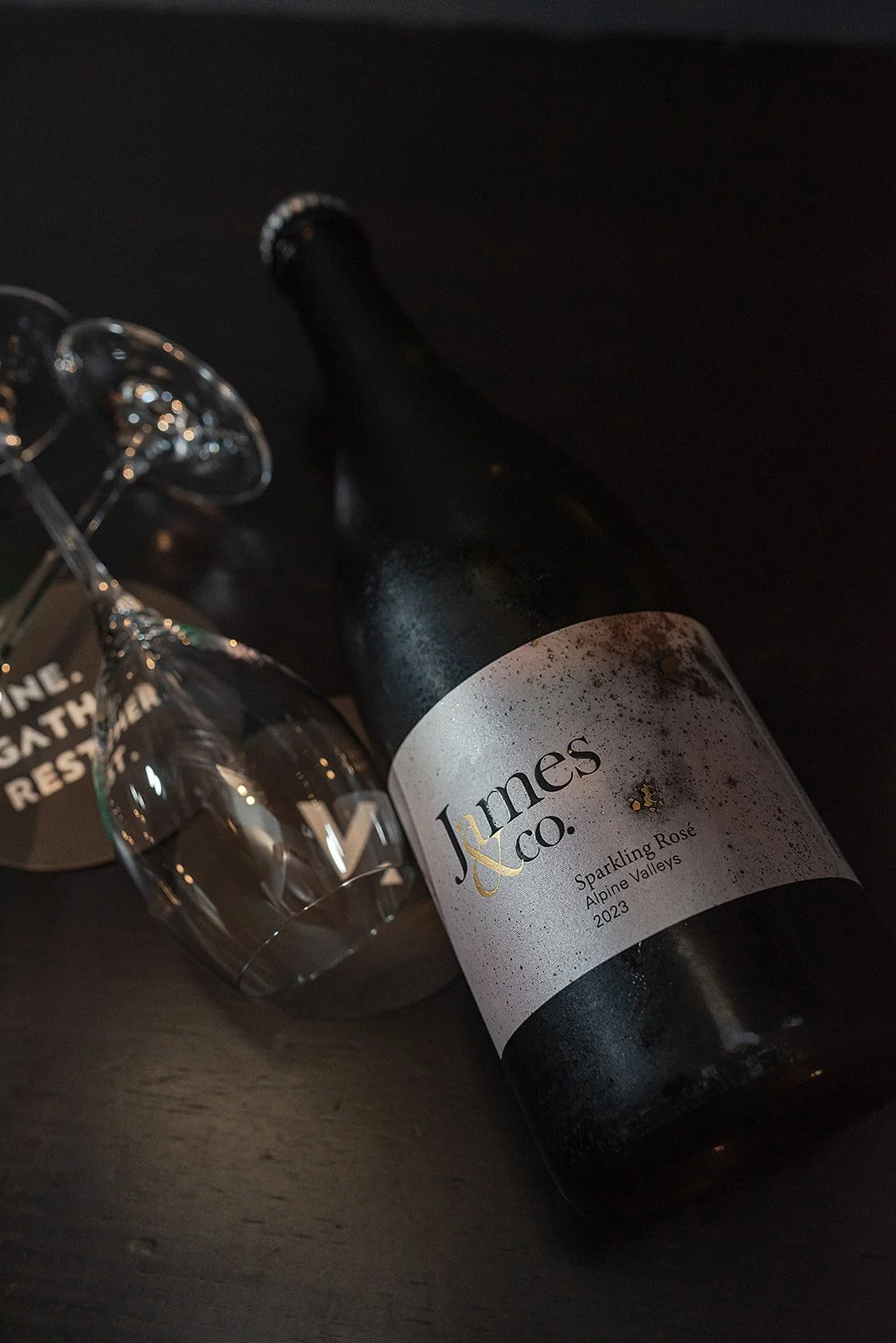 A bottle of James & Co. Sparkling Rosé wine from Alpiine Valleys, 2023, lying on a dark surface with a wine glass nearby.