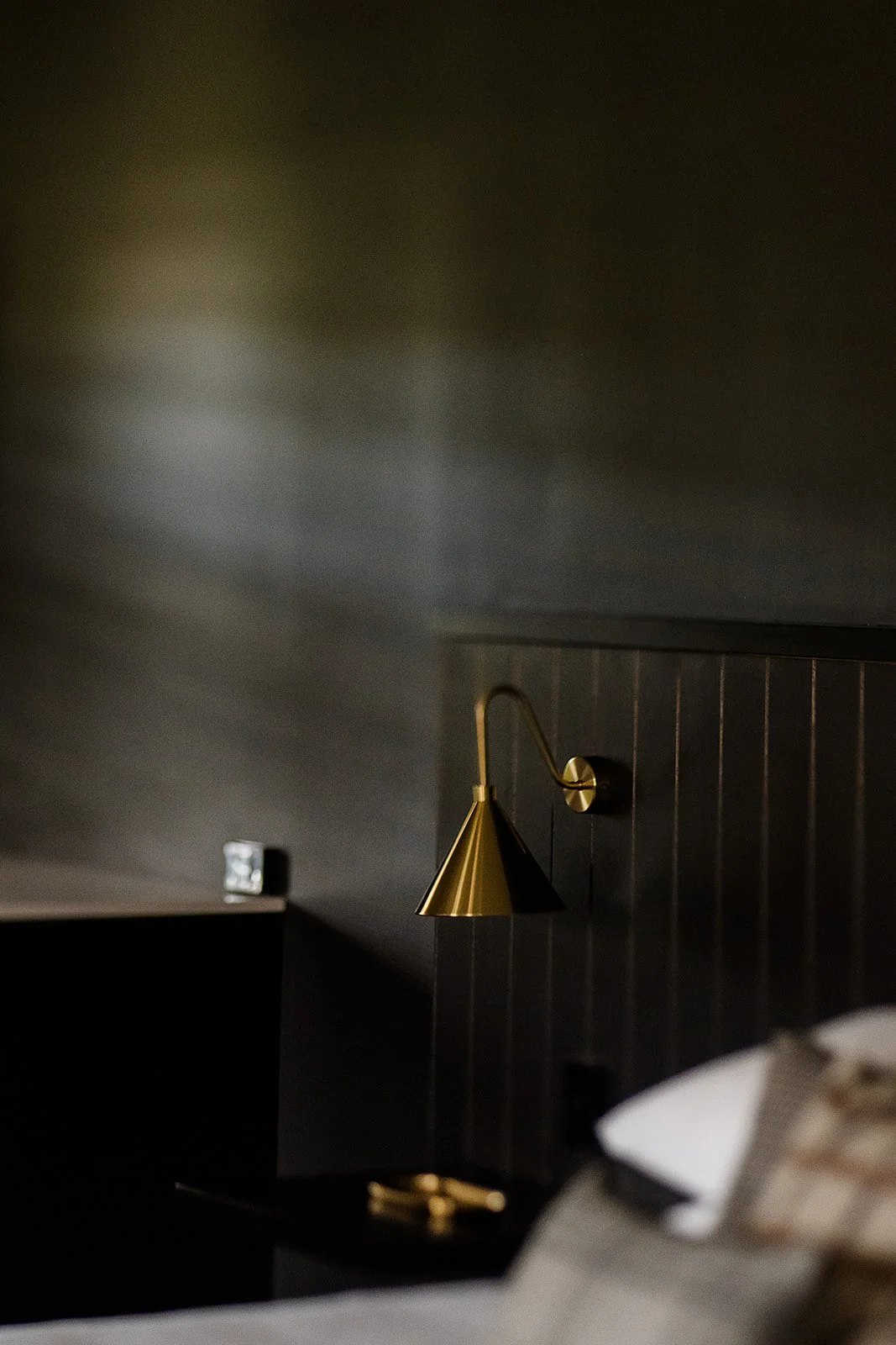 Close-up of a modern gold-colored bedside lamp mounted on a dark wood-paneled wall in a bedroom.