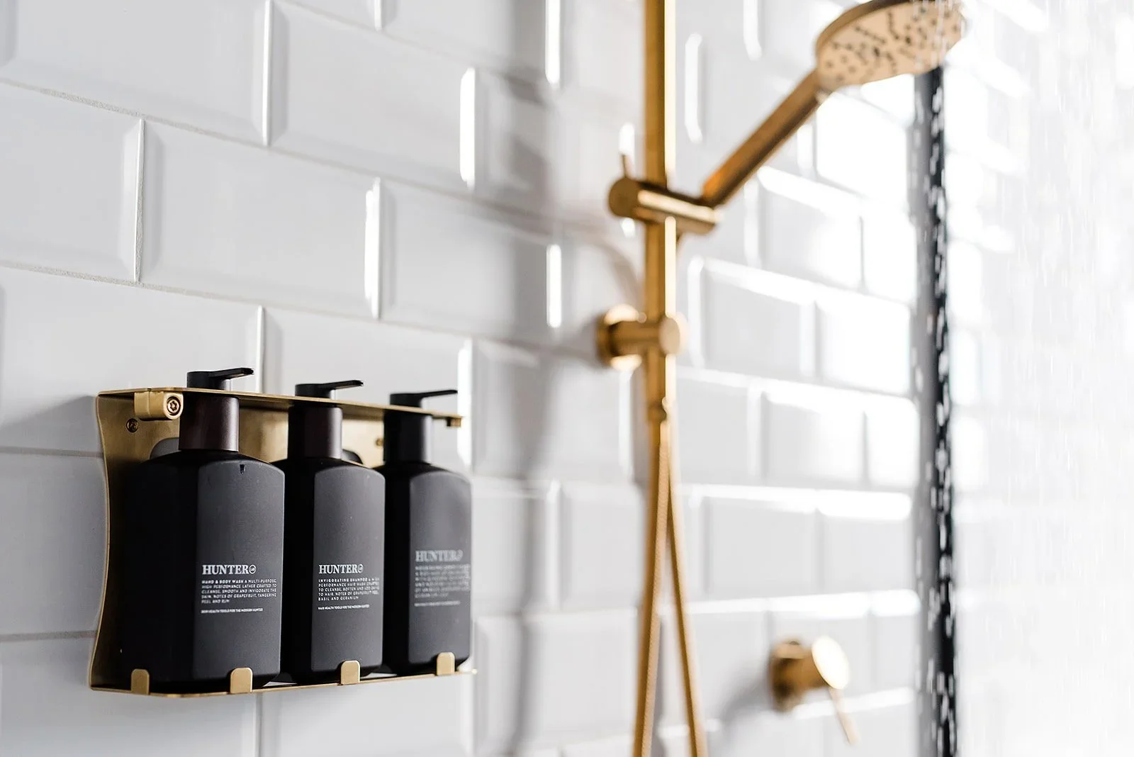 Bathroom wall with a mounted soap dispenser and shower fixtures. The soap dispenser is black with gold accents and labeled "HUNTER." The wall has white subway tiles.